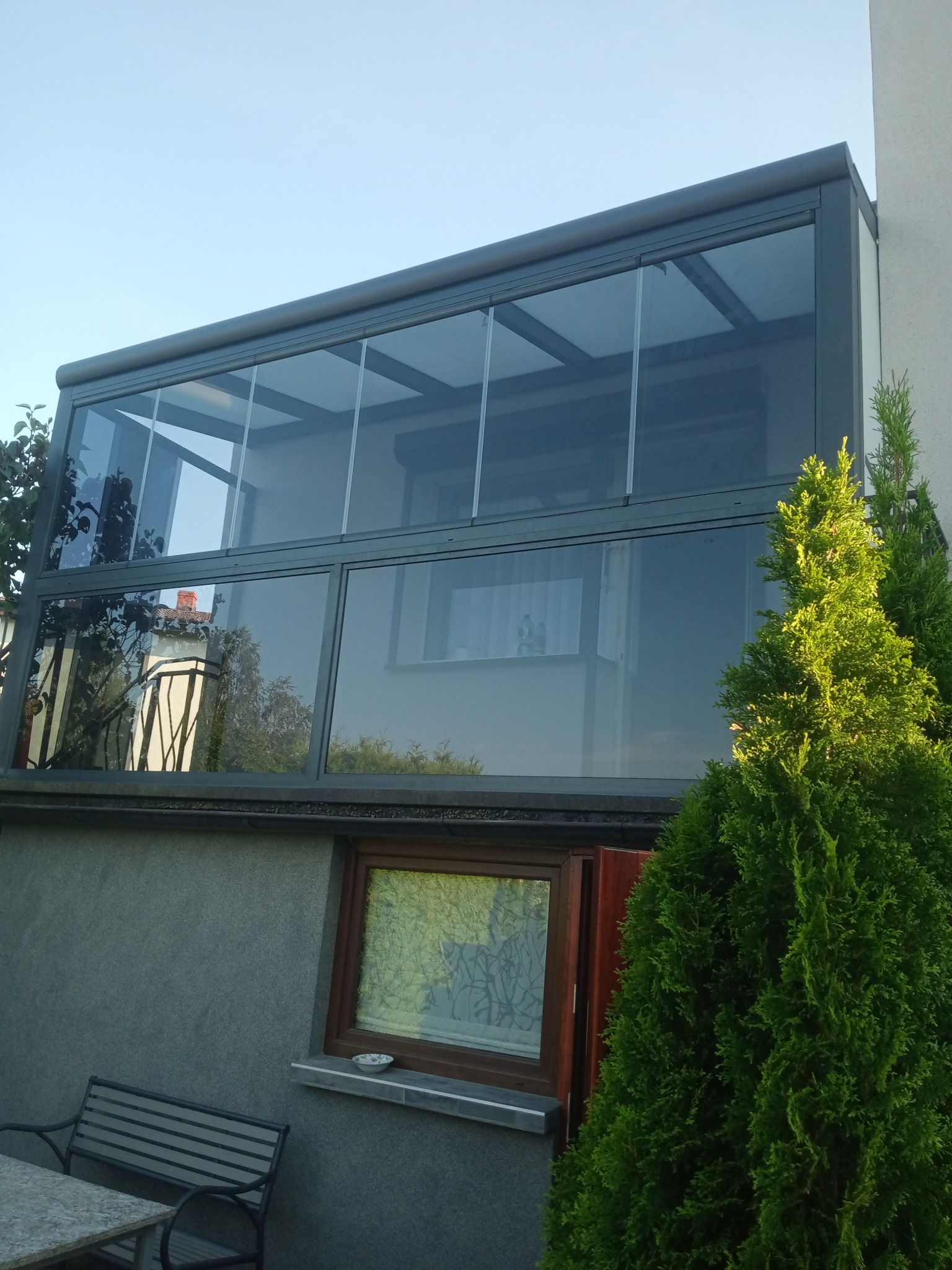 Zewnętrzny widok przeszklonej zabudowy balkonu z szarymi ramami, z roletami przeciwsłonecznymi częściowo opuszczonymi, z widocznymi odbiciami otoczenia w szybach i fragmentem iglaka w prawym dolnym...