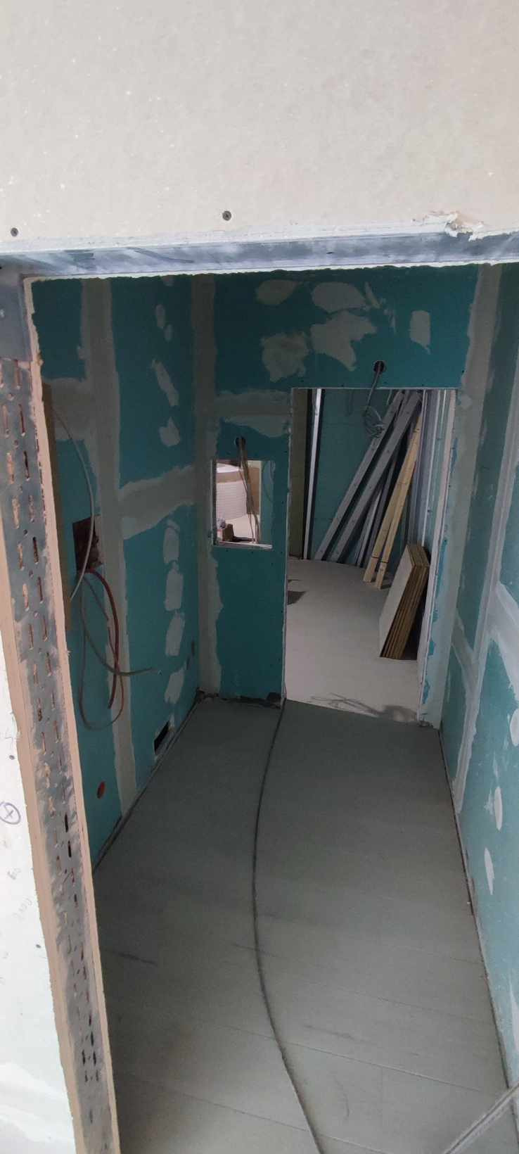 Wnętrze w trakcie prac wykończeniowych: ściany z płyt gipsowo-kartonowych z widocznymi łączeniami przed położeniem warstwy wykończeniowej, szare wylewki podłogowe, widoczne przewody elektryczne.