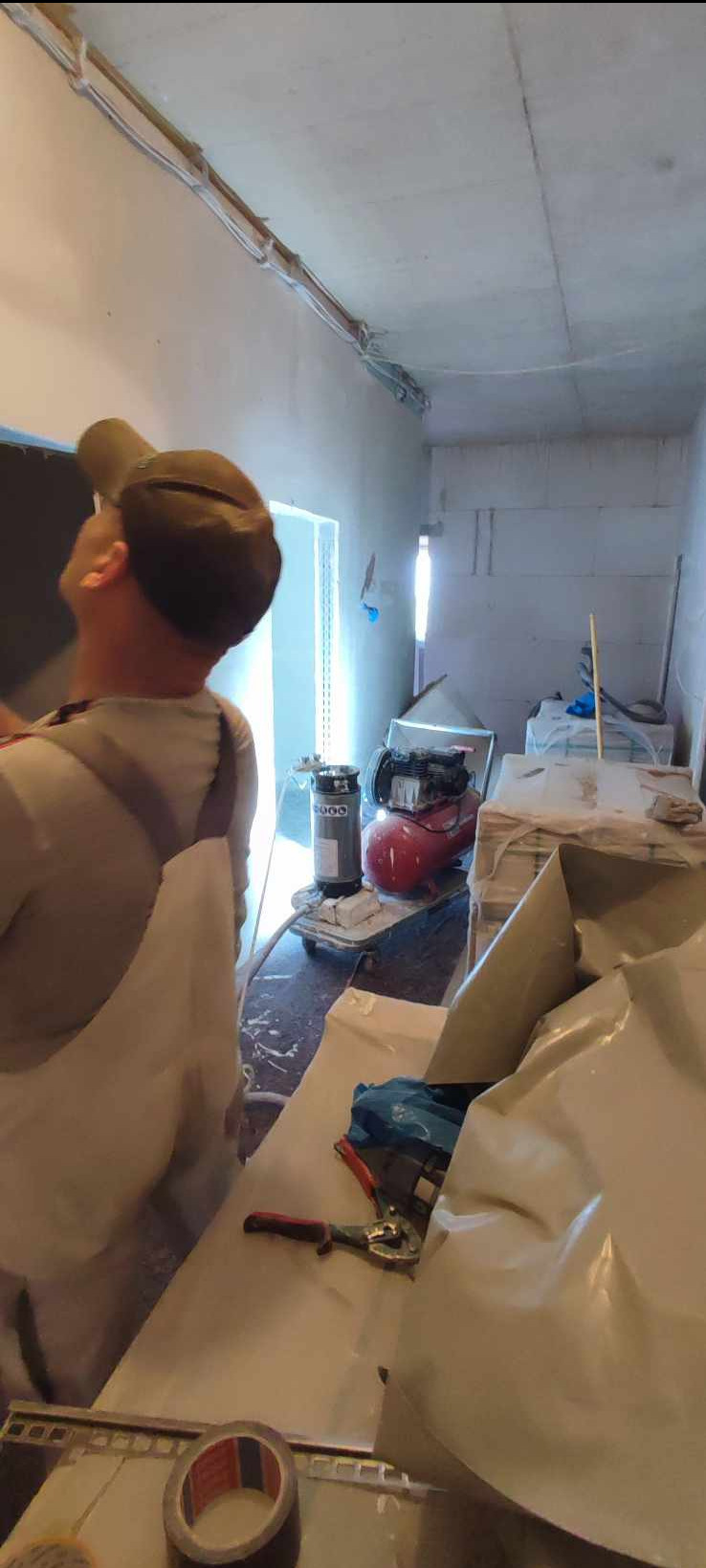 Pracownik w kombinezonie spogląda w górę na świeżo nałożoną warstwę gładzi na ścianie, widoczny kompresor i narzędzia do szpachlowania, przygotowane do dalszych prac wykończeniowych.