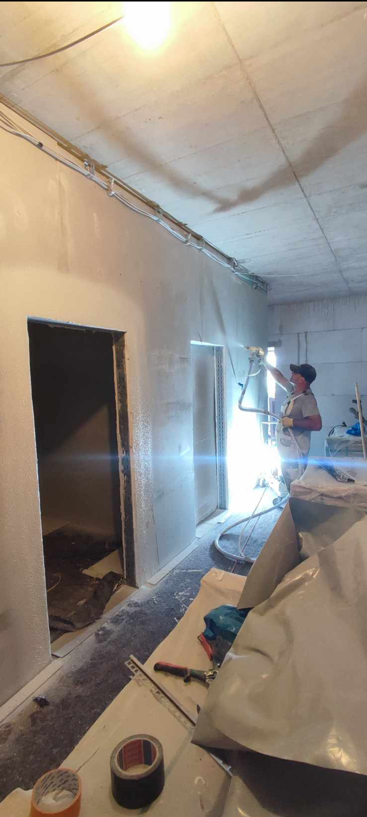 Pracownik w kombinezonie ochronnym aplikuje natryskowo masę na ściany wewnątrz budynku, widoczne przewody elektryczne pod sufitem, narzędzia i folie ochronne na podłodze.