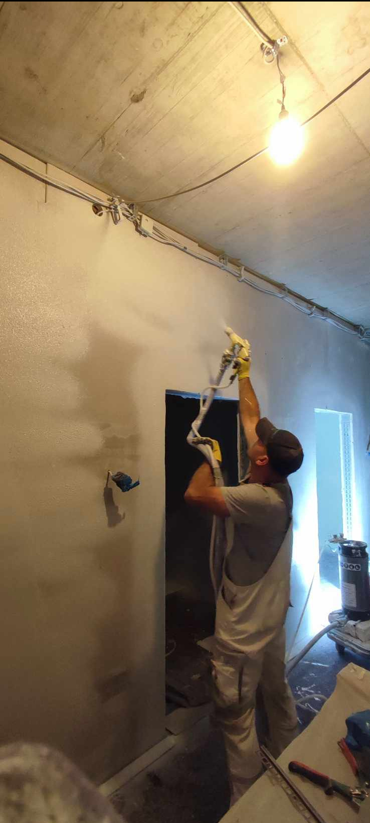 Pracownik w kombinezonie i rękawicach ochronnych aplikuje natryskowo gładź na ścianę w surowym pomieszczeniu z widoczną instalacją elektryczną pod sufitem i wiszącą żarówką.
