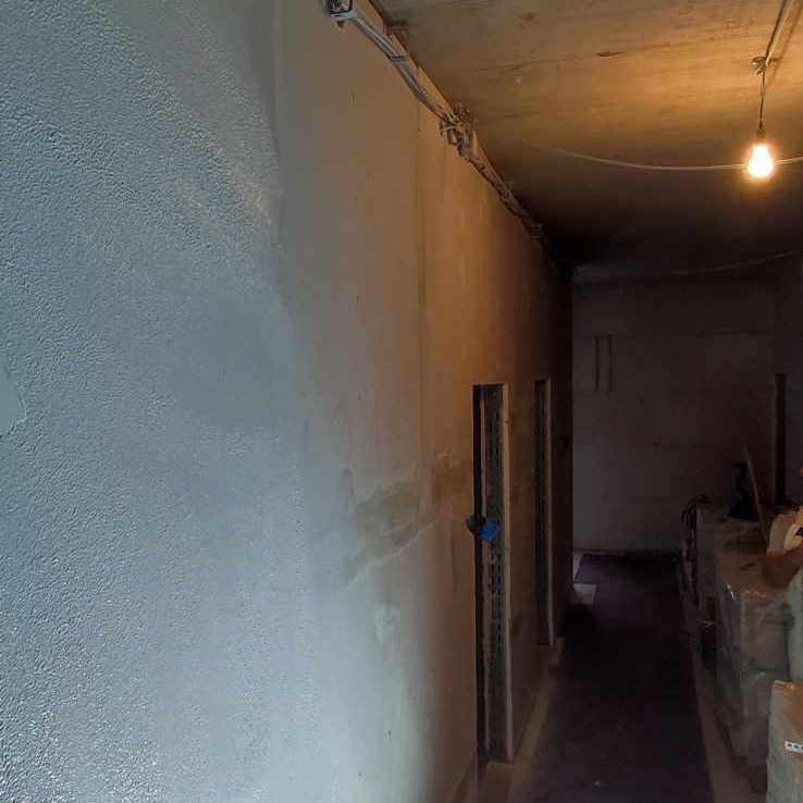 Korytarz w trakcie szpachlowania: ściana z widoczną fakturą i miejscami świeżo nałożonej gładzi, surowy sufit z instalacją elektryczną, widoczne ościeżnice drzwiowe.