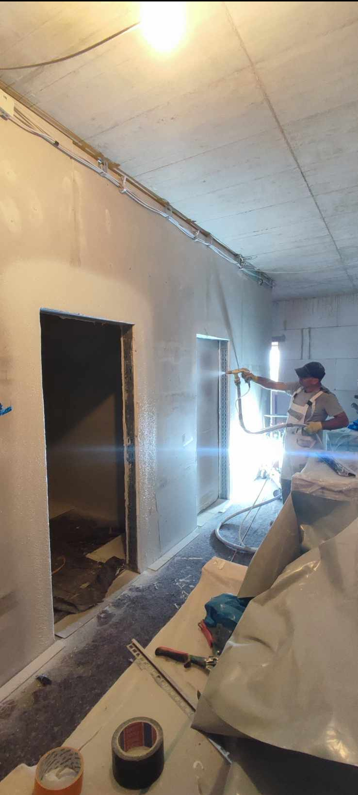 Pracownik w kombinezonie ochronnym aplikuje natryskowo masę na ścianę w surowym wnętrzu, widoczne przewody elektryczne pod sufitem, przygotowane narzędzia i folie ochronne na podłodze.