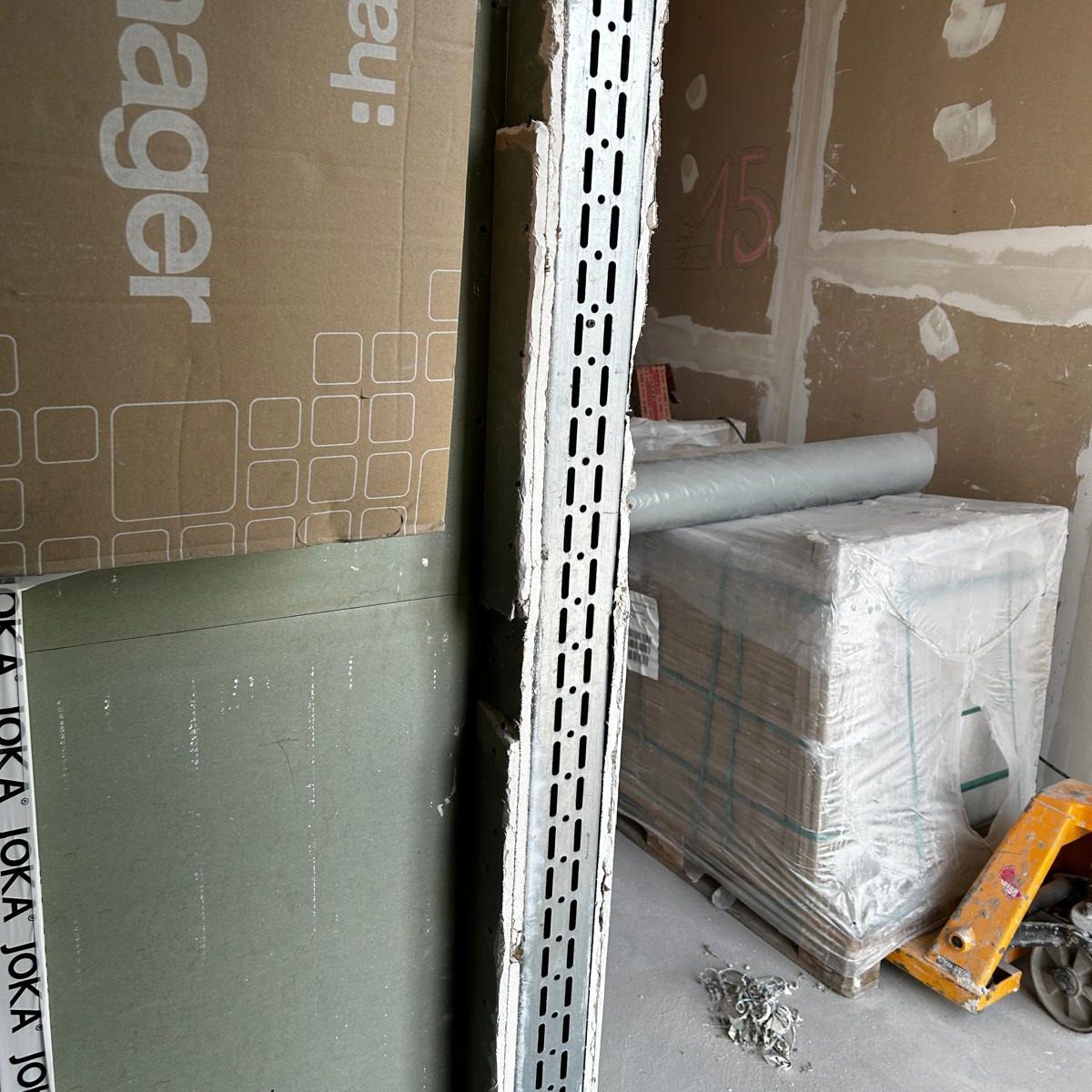 Stos płyt gipsowo-kartonowych JOKA, profil metalowy z perforacją, rolka folii i paleta z materiałami budowlanymi na tle ściany przygotowanej do wykończenia.