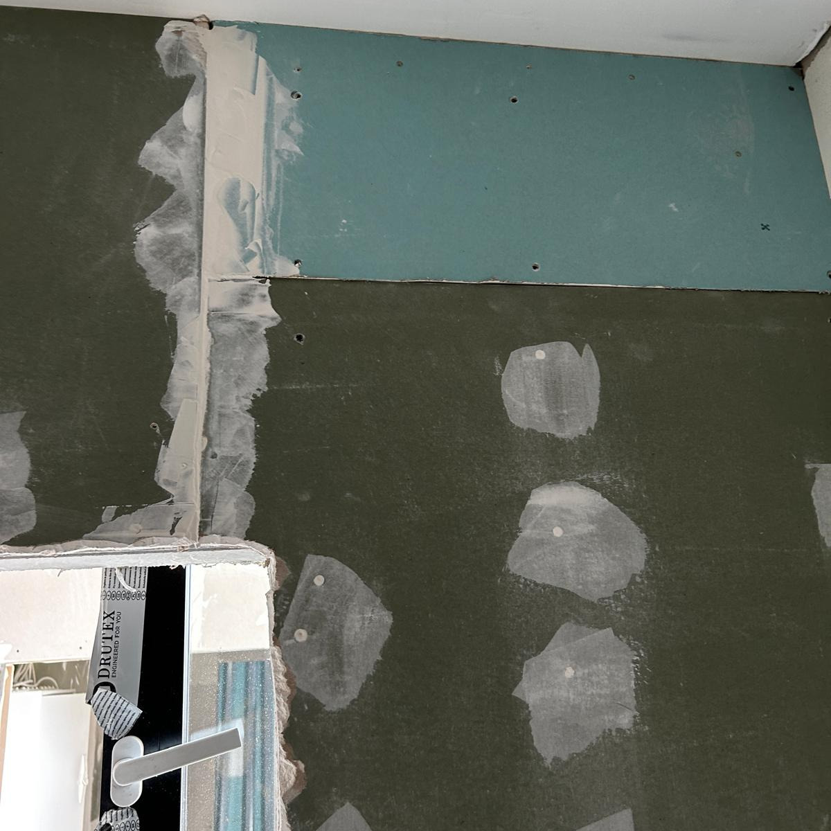 Przygotowanie ściany z płyt gipsowo-kartonowych do dalszych prac wykończeniowych, widoczne łączenia płyt wypełnione masą szpachlową oraz punktowe miejsca po wkrętach również zaszpachlowane, dolna...