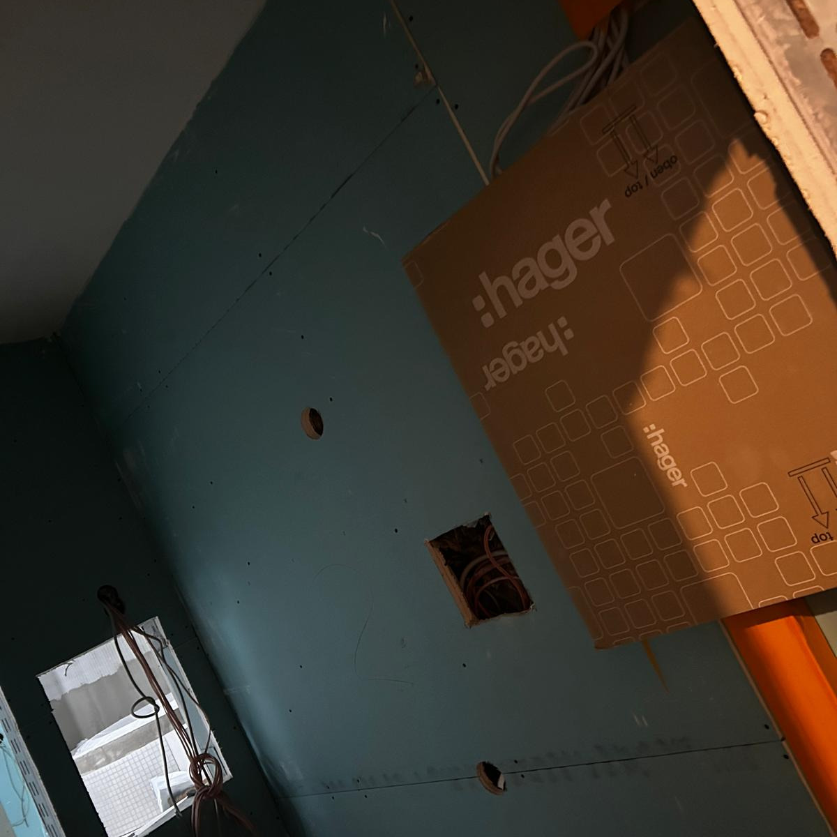 Wnętrze w trakcie prac wykończeniowych: ściany pokryte płytami gipsowo-kartonowymi z widocznymi otworami na instalacje elektryczne i puszką instalacyjną Hager, w tle widoczne okno z przewodami.