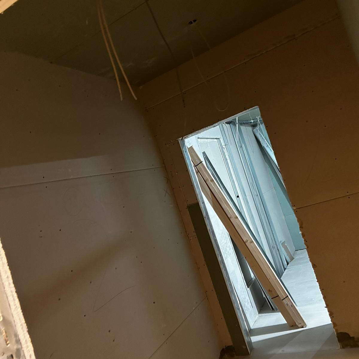 Wnętrze w trakcie prac wykończeniowych: ściany pokryte płytami gipsowo-kartonowymi, widoczne przewody elektryczne i drewniana rozpórka w otworze drzwiowym.