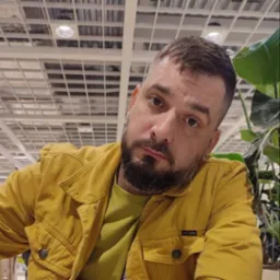 Portret mężczyzny z brodą w żółtej kurtce na tle sufitu z kratownicą i rośliny doniczkowej.