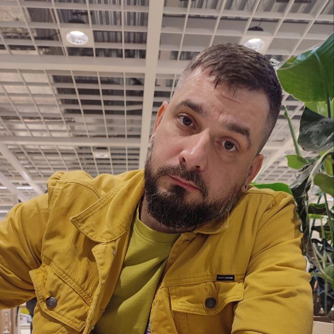 Portret mężczyzny z brodą w żółtej kurtce na tle sufitu z kratownicą i rośliny doniczkowej.