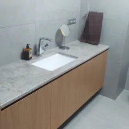 Nowoczesna łazienka z umywalką wpuszczoną w blat z szarego kamienia, drewniane szafki pod umywalką, minimalistyczna bateria, okrągłe lusterko kosmetyczne i szary ręcznik na tle szarych płytek.