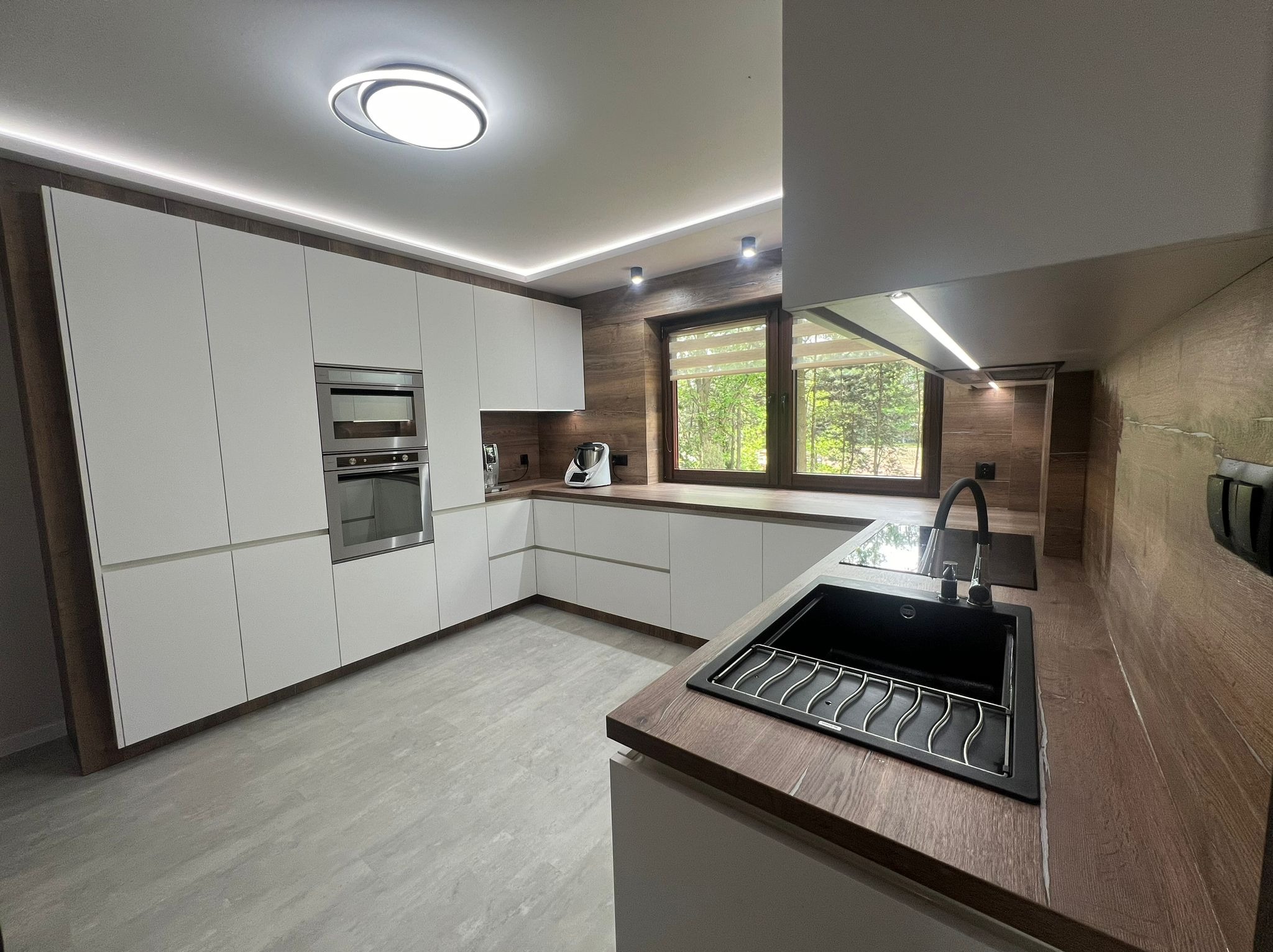 Nowoczesna kuchnia z białymi frontami szafek, drewnianym blatem i okładziną ścienną, czarnym zlewozmywakiem z ociekaczem i widokiem na zieleń za oknem.