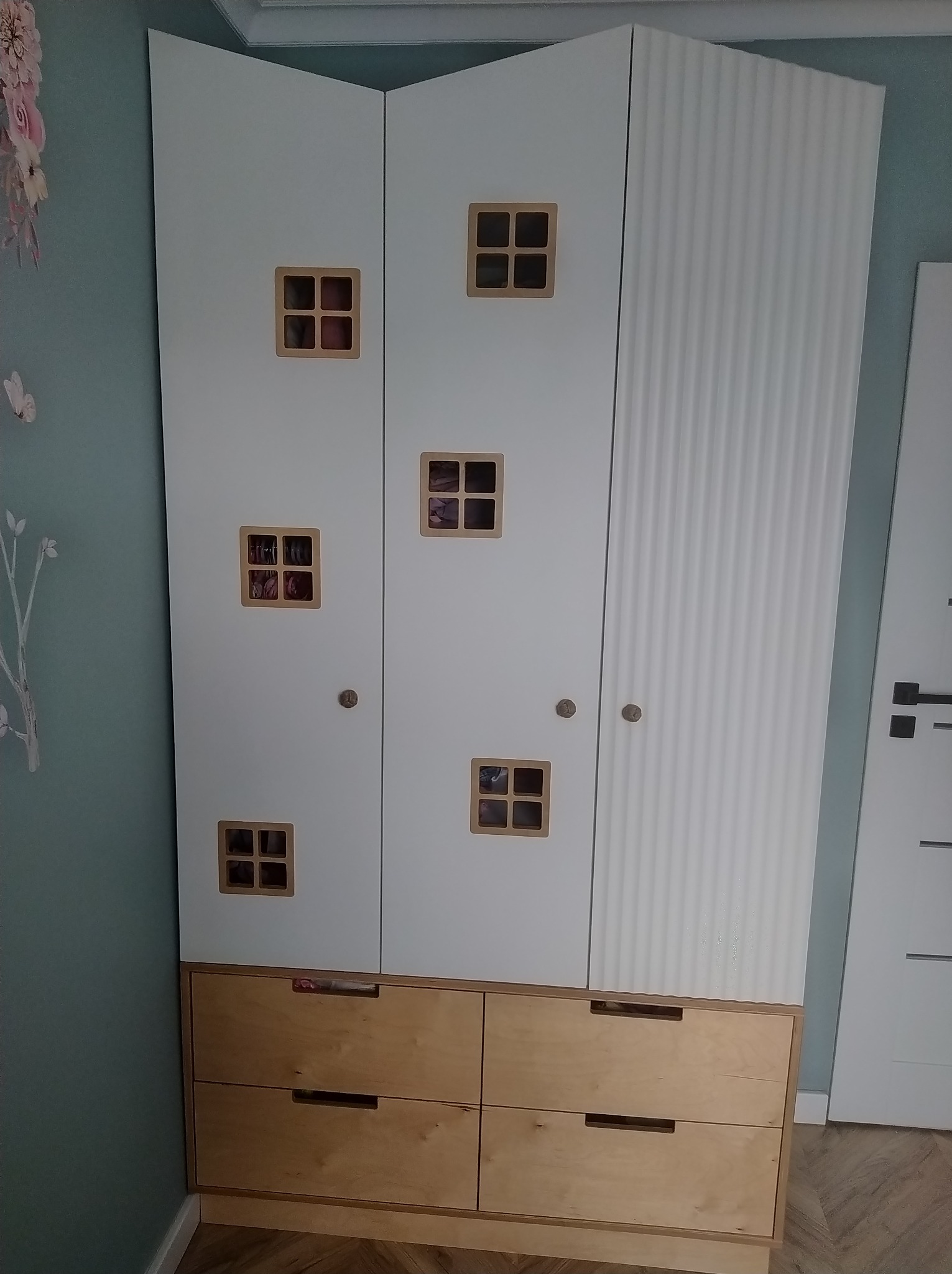 Biała szafa z drewnianymi szufladami i kwadratowymi okienkami, przypominająca domek dla lalek, z dekoracyjną ścianą w tle.