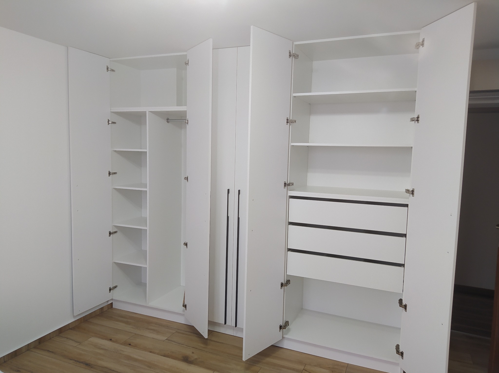 Biała szafa wnękowa z otwartymi drzwiami, widoczne półki i szuflady z czarnymi akcentami, parkiet na podłodze.