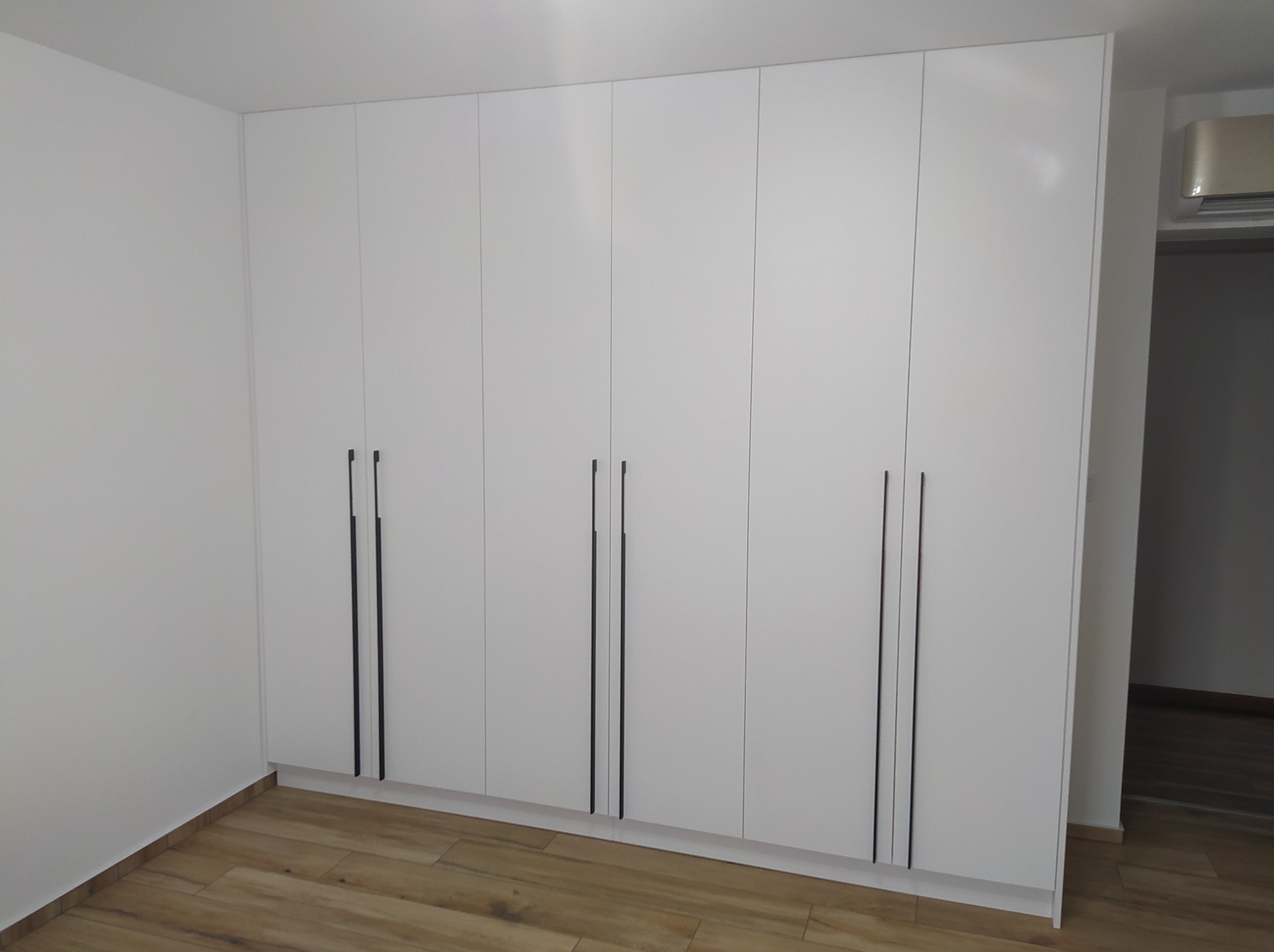 Biała zabudowa szafy z sześcioma drzwiami i minimalistycznymi, pionowymi, czarnymi uchwytami na tle parkietu i białych ścian.