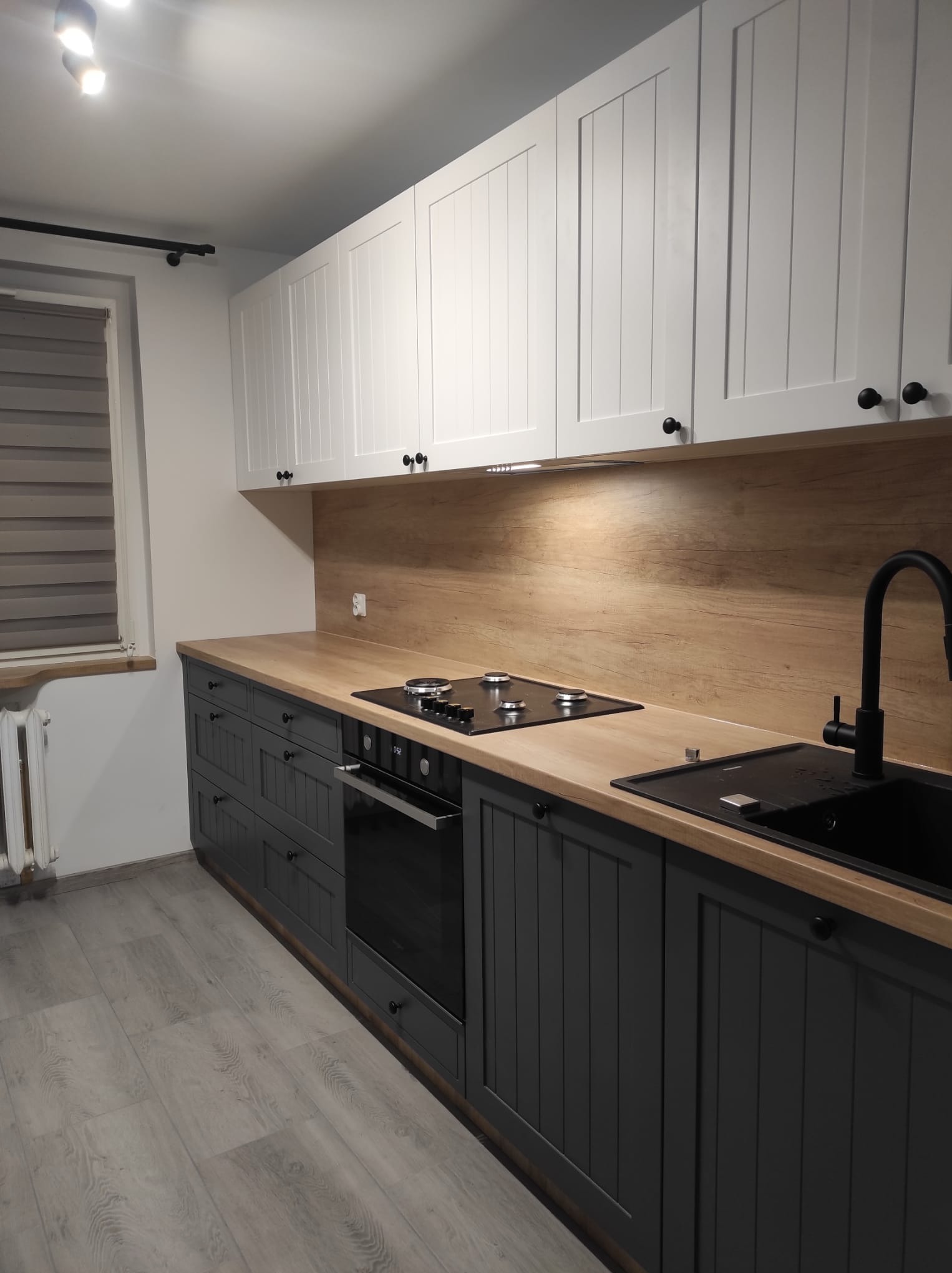 Nowoczesna kuchnia z białymi szafkami górnymi i ciemnoszarymi dolnymi, drewnianym blatem i czarną armaturą, widoczna płyta grzewcza i zlewozmywak, oświetlenie podszafkowe.