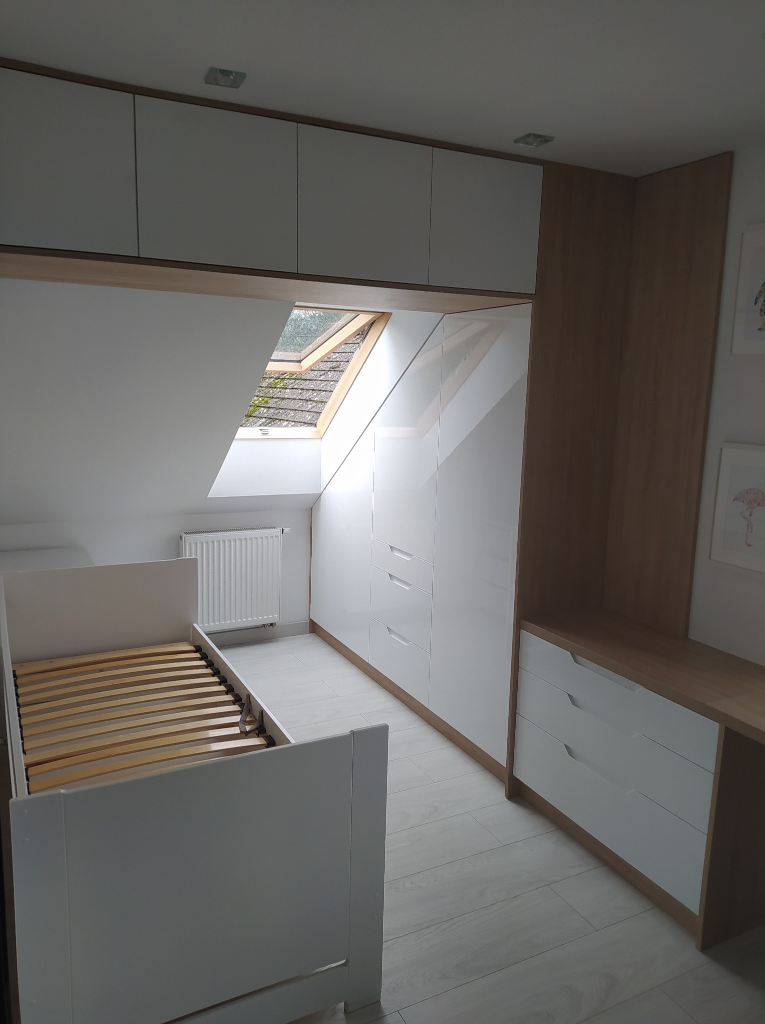 Pokój na poddaszu z białymi szafami wnękowymi z połyskiem, drewnianym blatem, łóżkiem bez materaca i oknem dachowym.