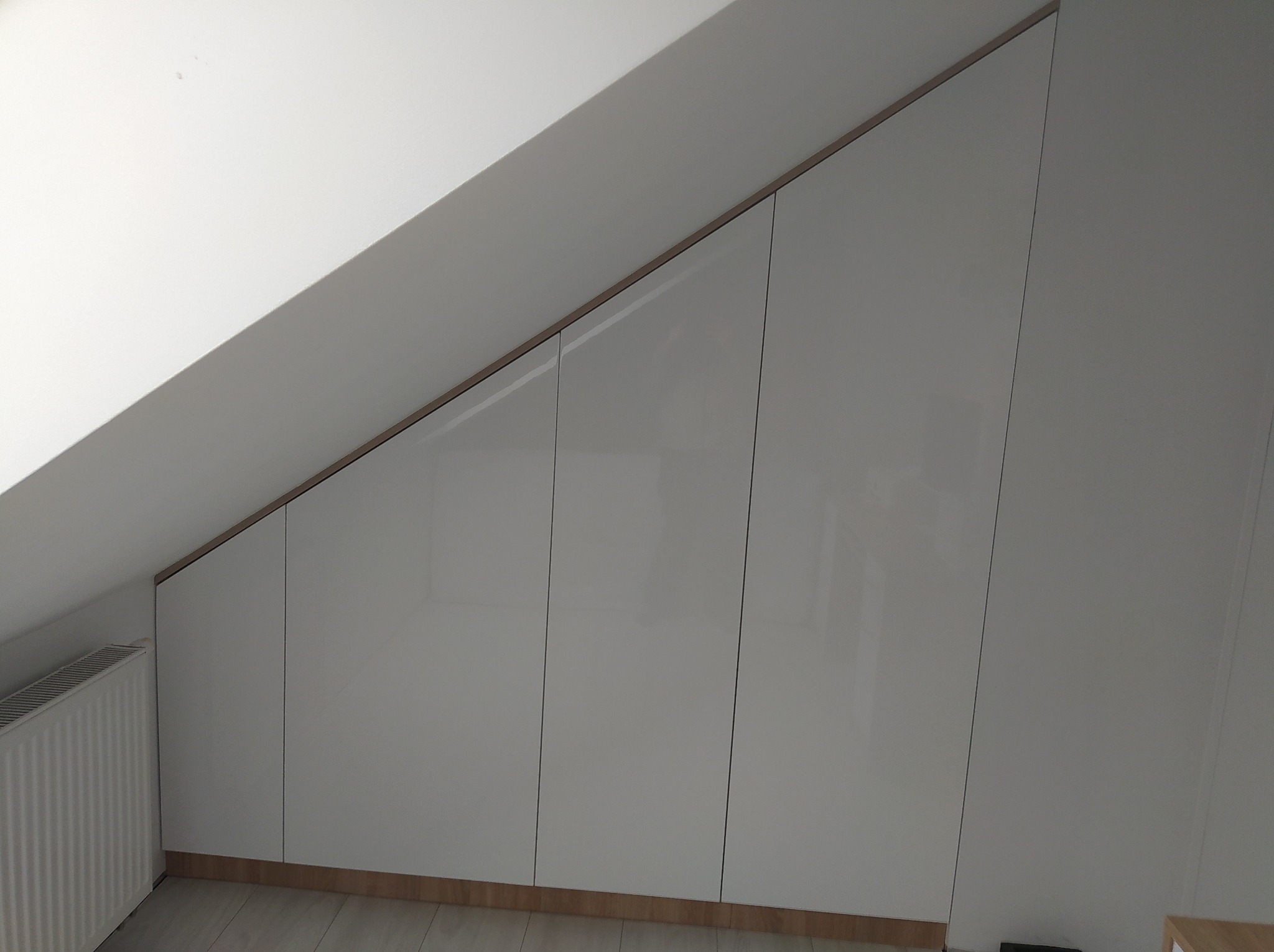 Białe, połyskujące szafy wnękowe z drewnianymi akcentami, dopasowane do skosu dachu na poddaszu, widoczny grzejnik.