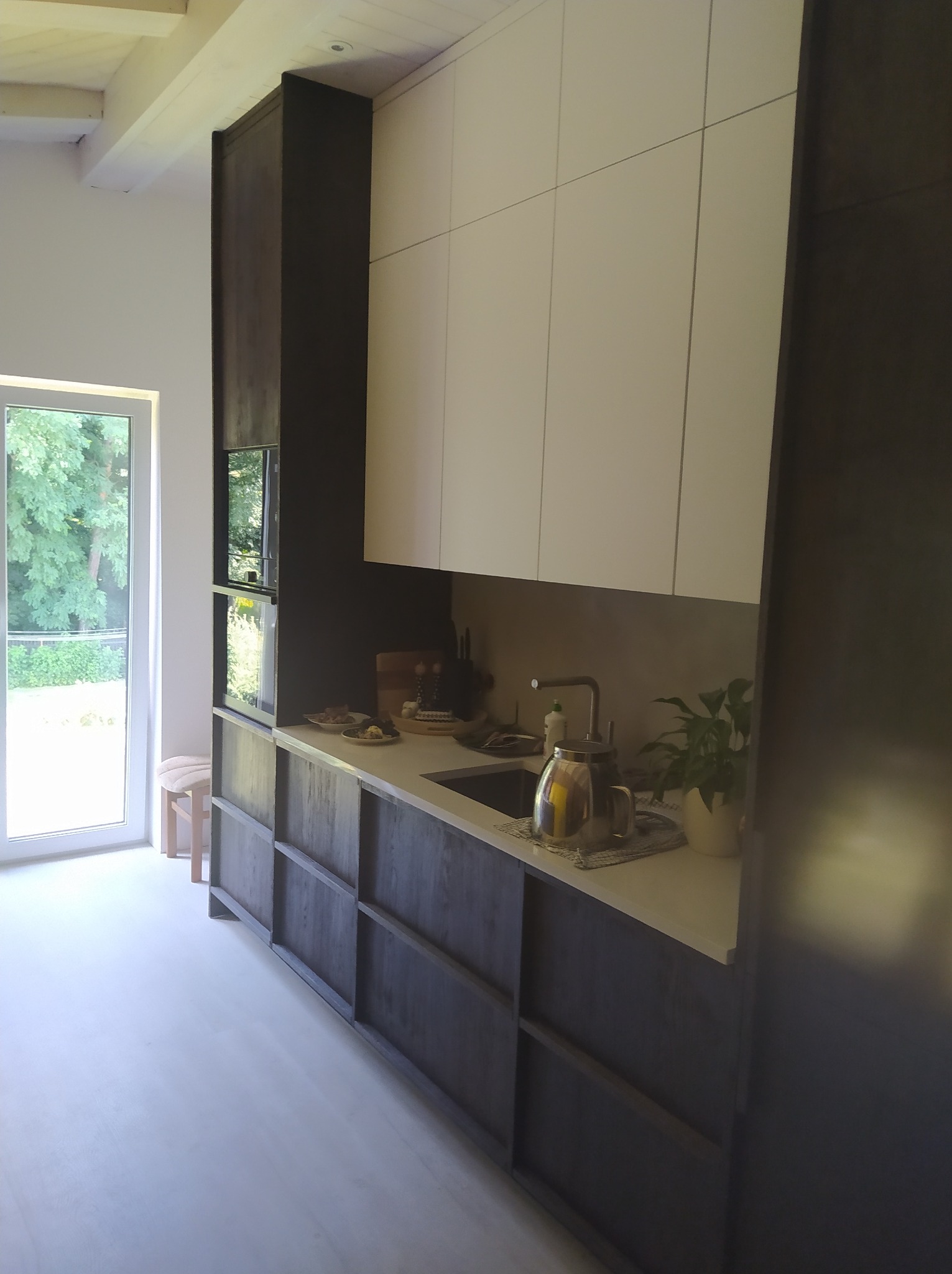 Nowoczesna kuchnia z ciemnymi, drewnianymi frontami szafek dolnych i białymi szafkami górnymi, jasny blat, widok na ogród przez duże okno.