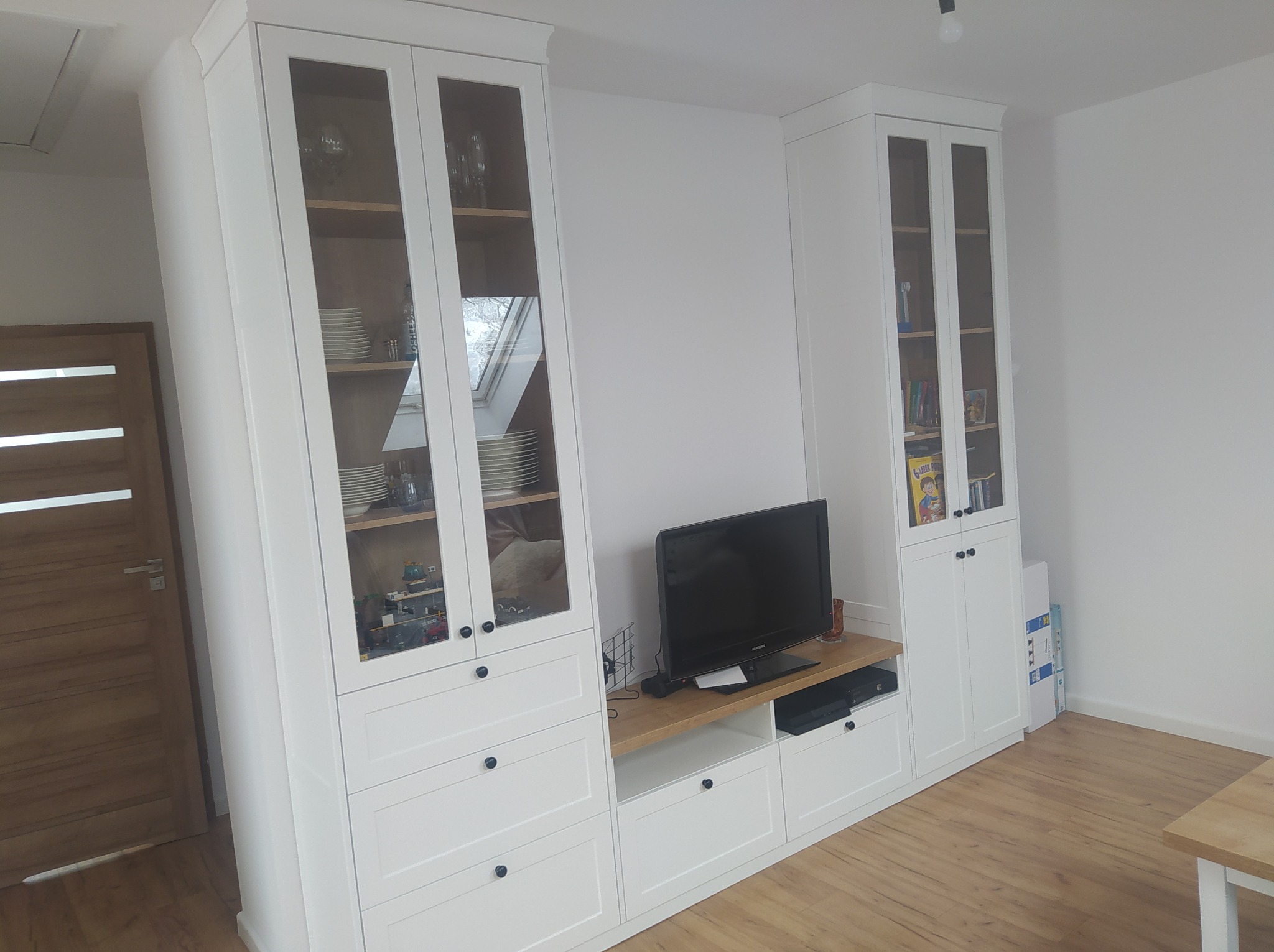 Białe meble modułowe w salonie, z przeszklonymi witrynami, drewnianymi półkami wewnętrznymi i szufladami, pod telewizorem drewniany blat, na półkach szkło, naczynia i dekoracje.