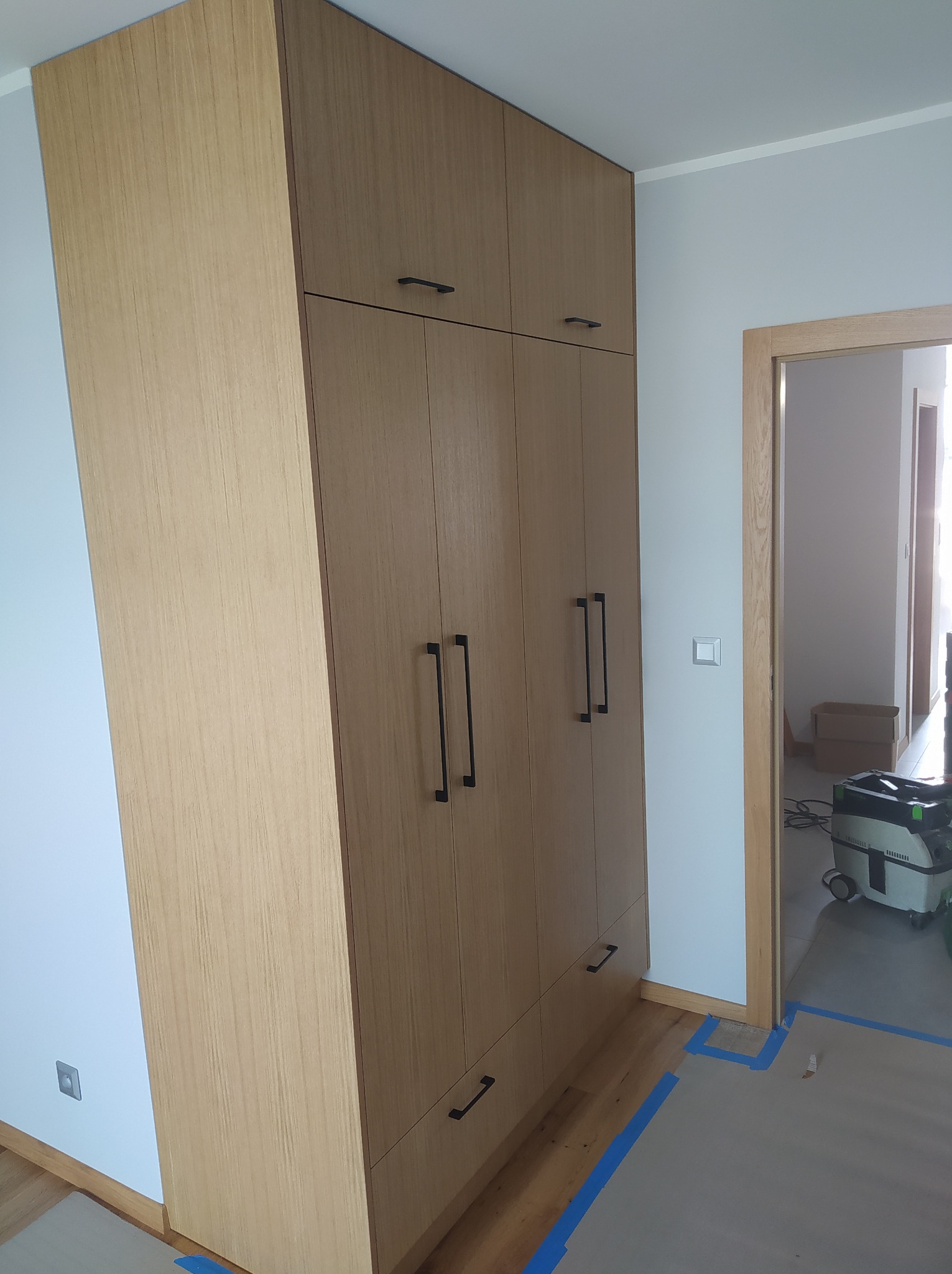 Wysoka szafa z jasnego drewna z czarnymi, prostokątnymi uchwytami, dopasowana do sufitu, na podłodze zabezpieczonej kartonem i niebieską taśmą, z odkurzaczem przemysłowym w tle.