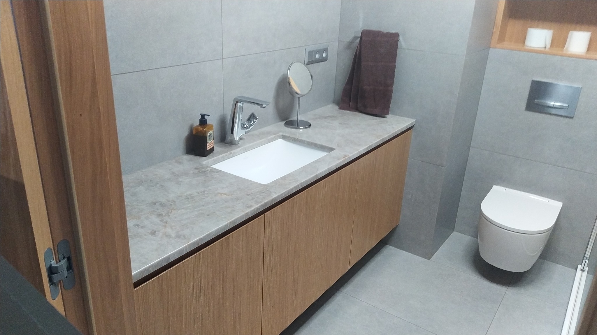 Nowoczesna łazienka z umywalką wpuszczoną w blat z szarego kamienia, drewniane szafki pod umywalką, minimalistyczna bateria, okrągłe lusterko kosmetyczne i szary ręcznik na tle szarych płytek.