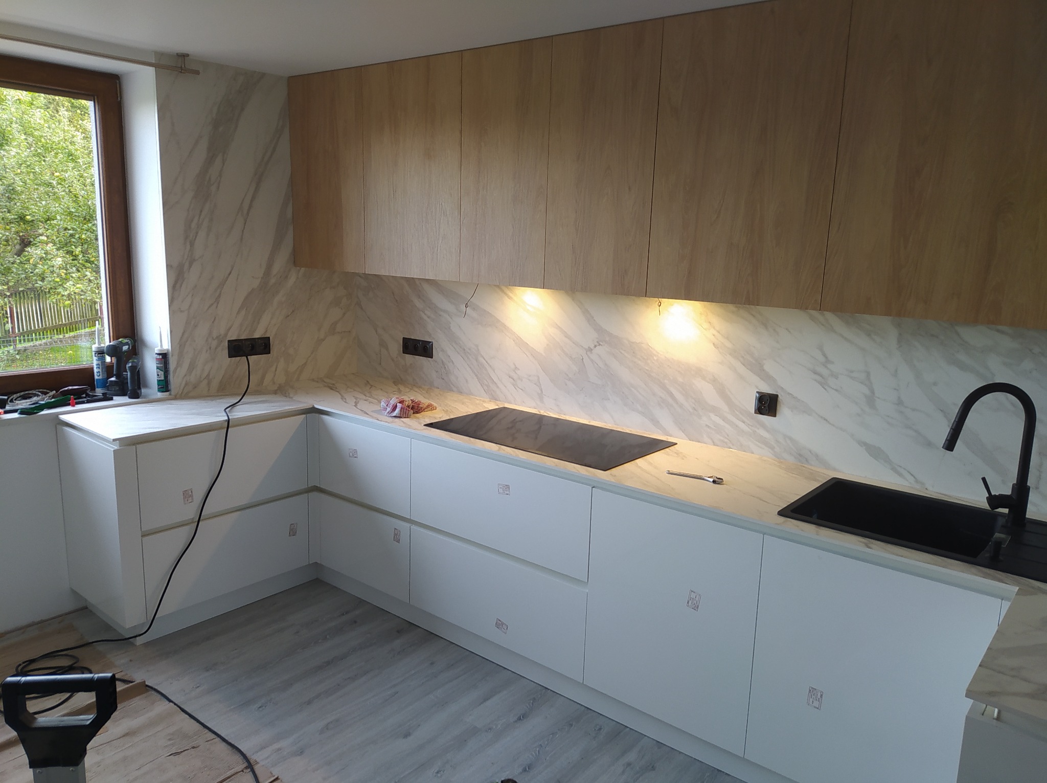 Nowoczesna kuchnia z białymi szafkami dolnymi i drewnopodobnymi górnymi, blat z marmurowym wzorem, czarny zlew i płyta indukcyjna, widoczne narzędzia i akcesoria na blacie, podświetlenie LED...