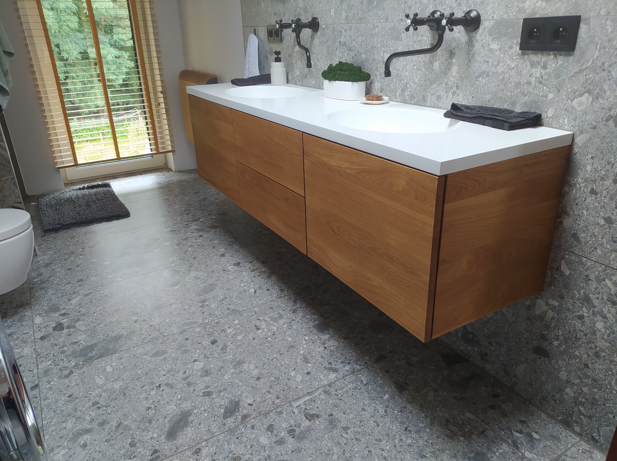 Nowoczesna łazienka w Pruchna z podwójną umywalką na drewnianej szafce wiszącej, biały blat, czarne baterie retro i szara, wzorzysta posadzka.