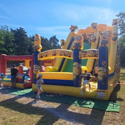 Kraina Smaków i Zabawy - Dzieci bawią się na dmuchanym zamku z motywem Minionków, ustawionym na trawie w słoneczny dzień. Widoczny również dmuchany plac zabaw z siatką w tle.