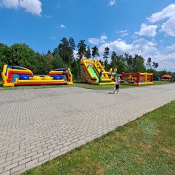 Kraina Smaków i Zabawy - Zestaw kolorowych dmuchanych atrakcji, w tym tor przeszkód i zjeżdżalnia z motywem Minionków, rozstawiony na utwardzonym placu w plenerze z drzewami w tle i osobą przechodzącą obok.