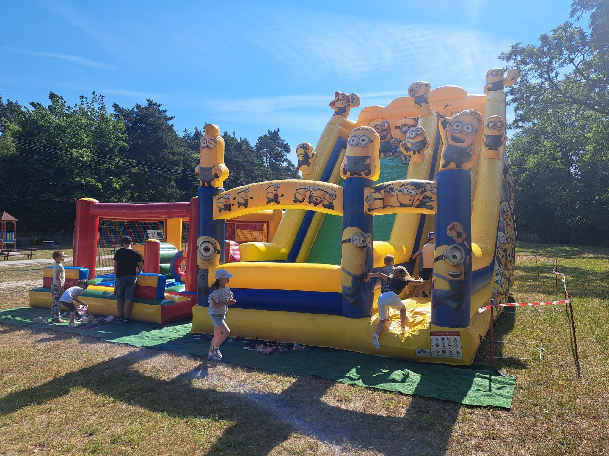 Dzieci bawią się na dmuchanym zamku z motywem Minionków, ustawionym na trawie w słoneczny dzień. Widoczny również dmuchany plac zabaw z siatką w tle.
