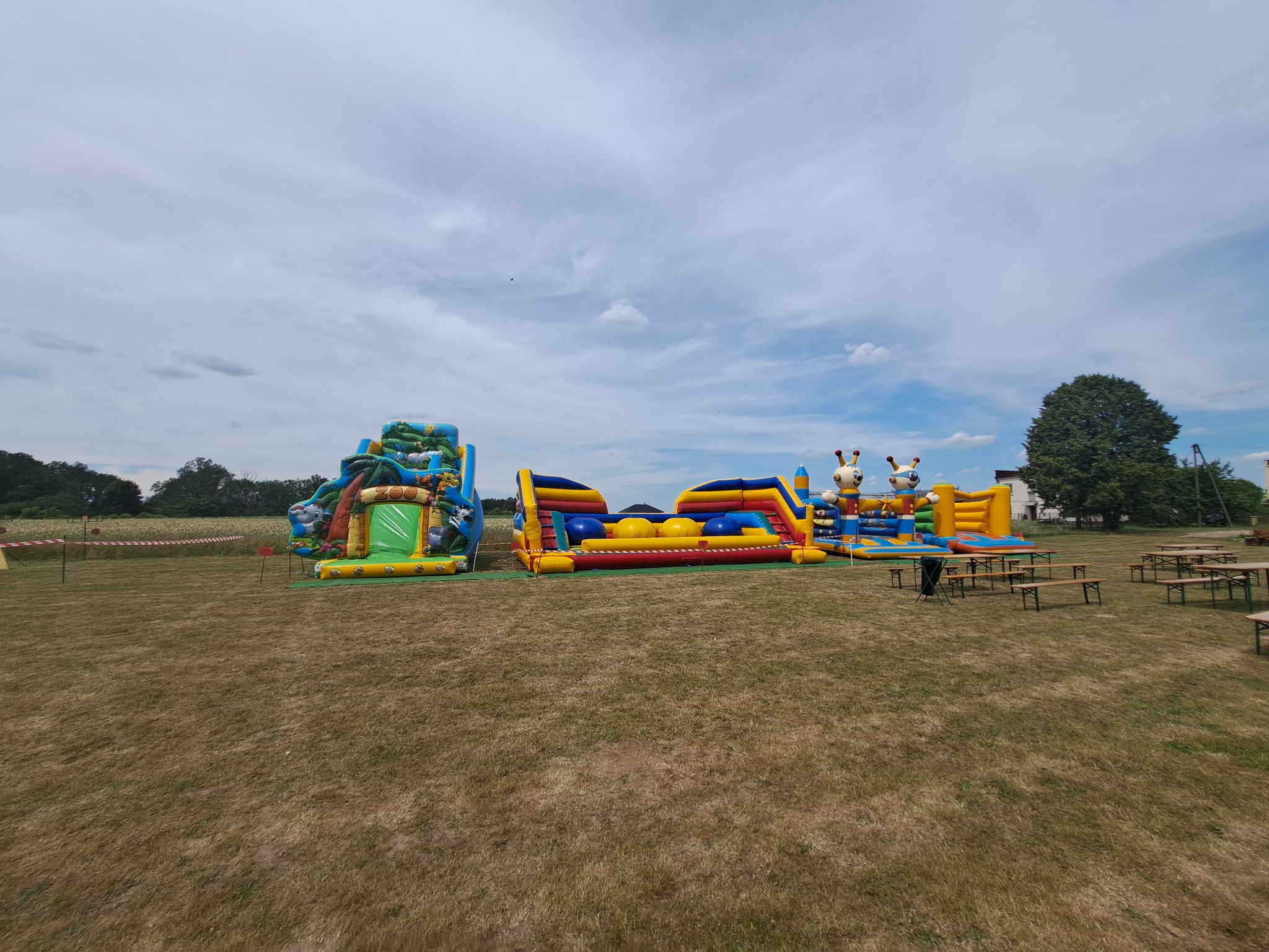 Kompleks dmuchańców w plenerze: zjeżdżalnia z motywem zoo, tor przeszkód i dmuchany plac zabaw z żyrafami, ustawione na trawiastym terenie pod błękitnym niebem z chmurami.