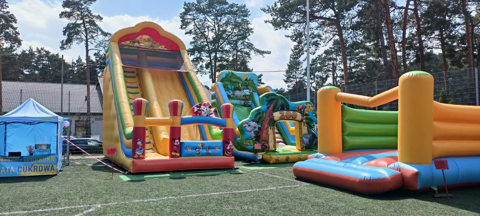 Kompleks dmuchańców z motywami z kreskówek i zoo, ustawiony na zielonej trawie, obok stoisko z watą cukrową i popcornem, w tle drzewa.
