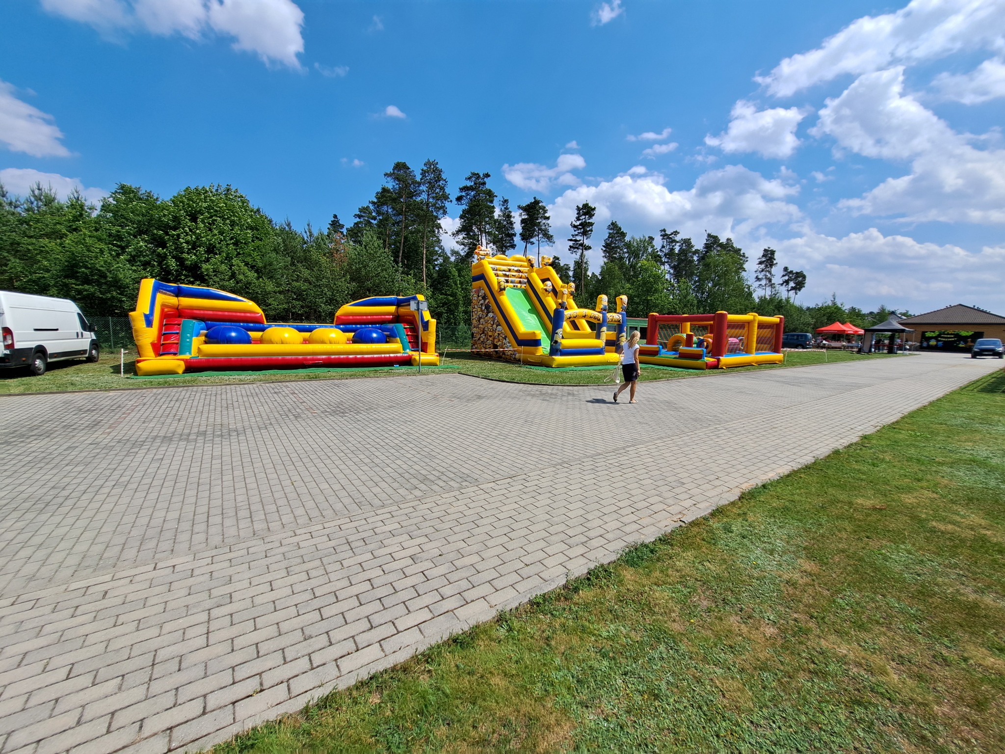 Zestaw kolorowych dmuchanych atrakcji, w tym tor przeszkód i zjeżdżalnia z motywem Minionków, rozstawiony na utwardzonym placu w plenerze z drzewami w tle i osobą przechodzącą obok.