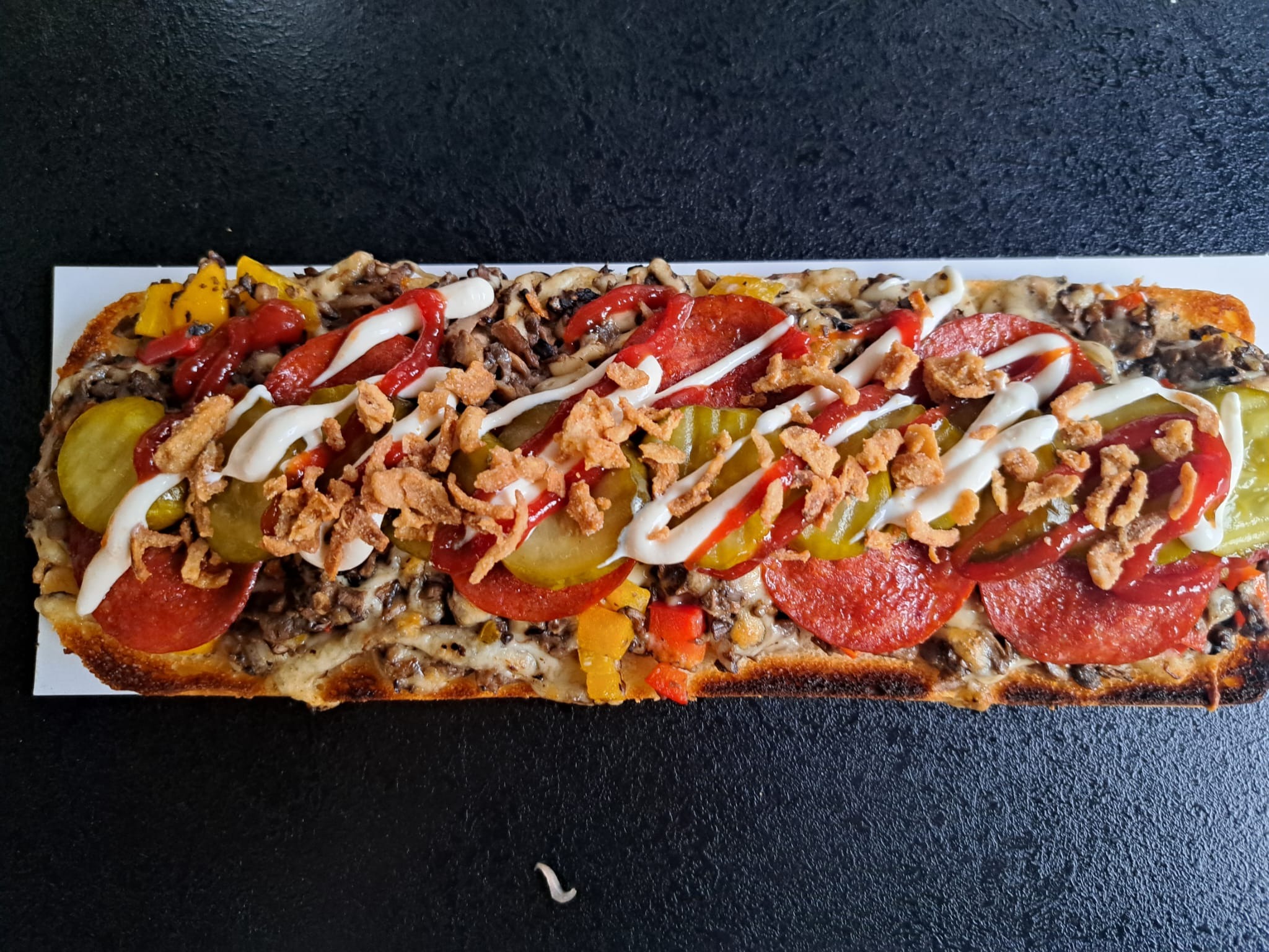 Długa zapiekanka na białej desce z pieczarkami, salami, ogórkami konserwowymi, papryką, sosem majonezowym, ketchupem i prażoną cebulką, ułożona na czarnym tle.