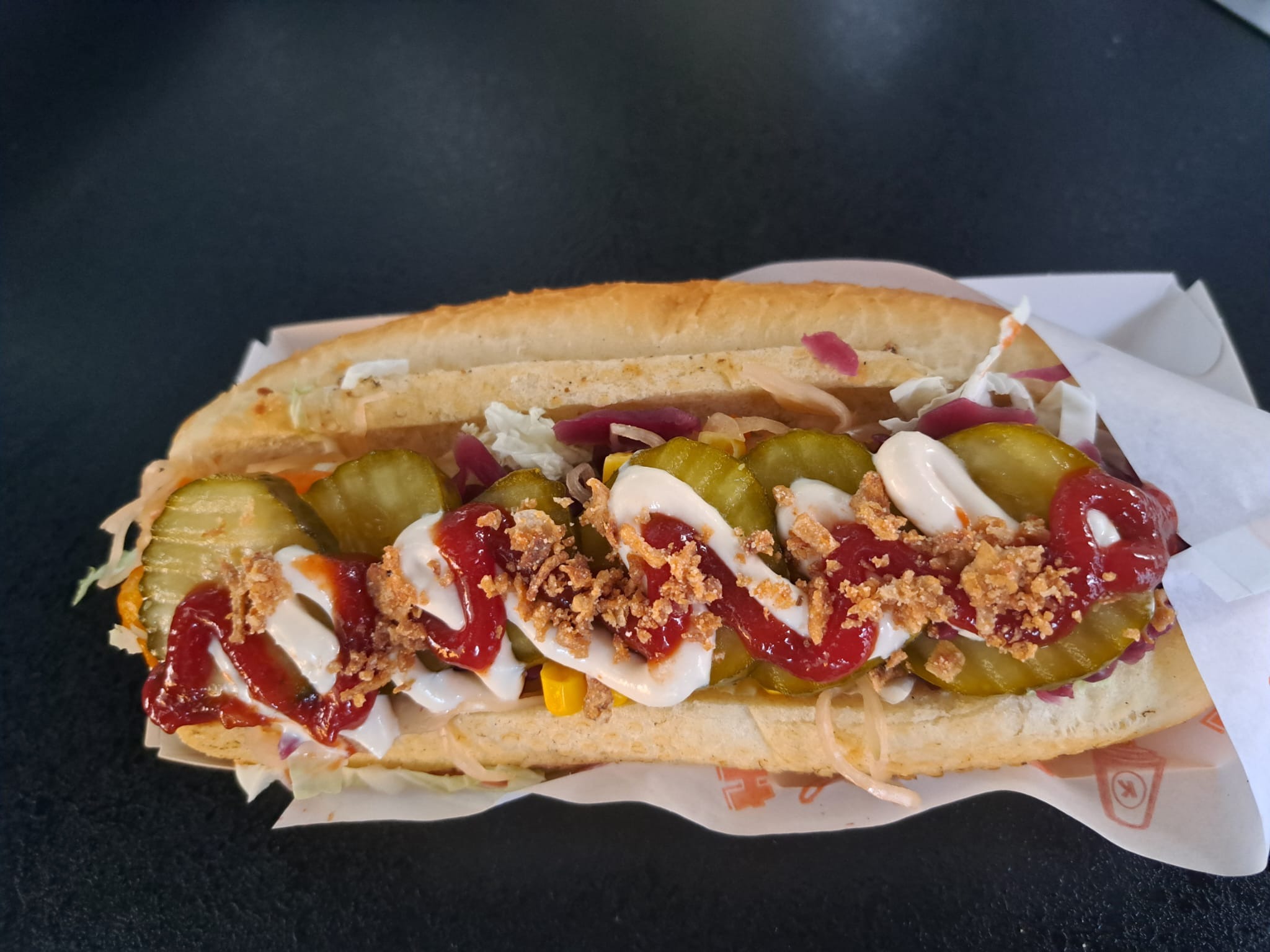 Długi hot dog z ogórkami konserwowymi, sosem majonezowym, ketchupem i prażoną cebulką, podany w papierowej tacce.