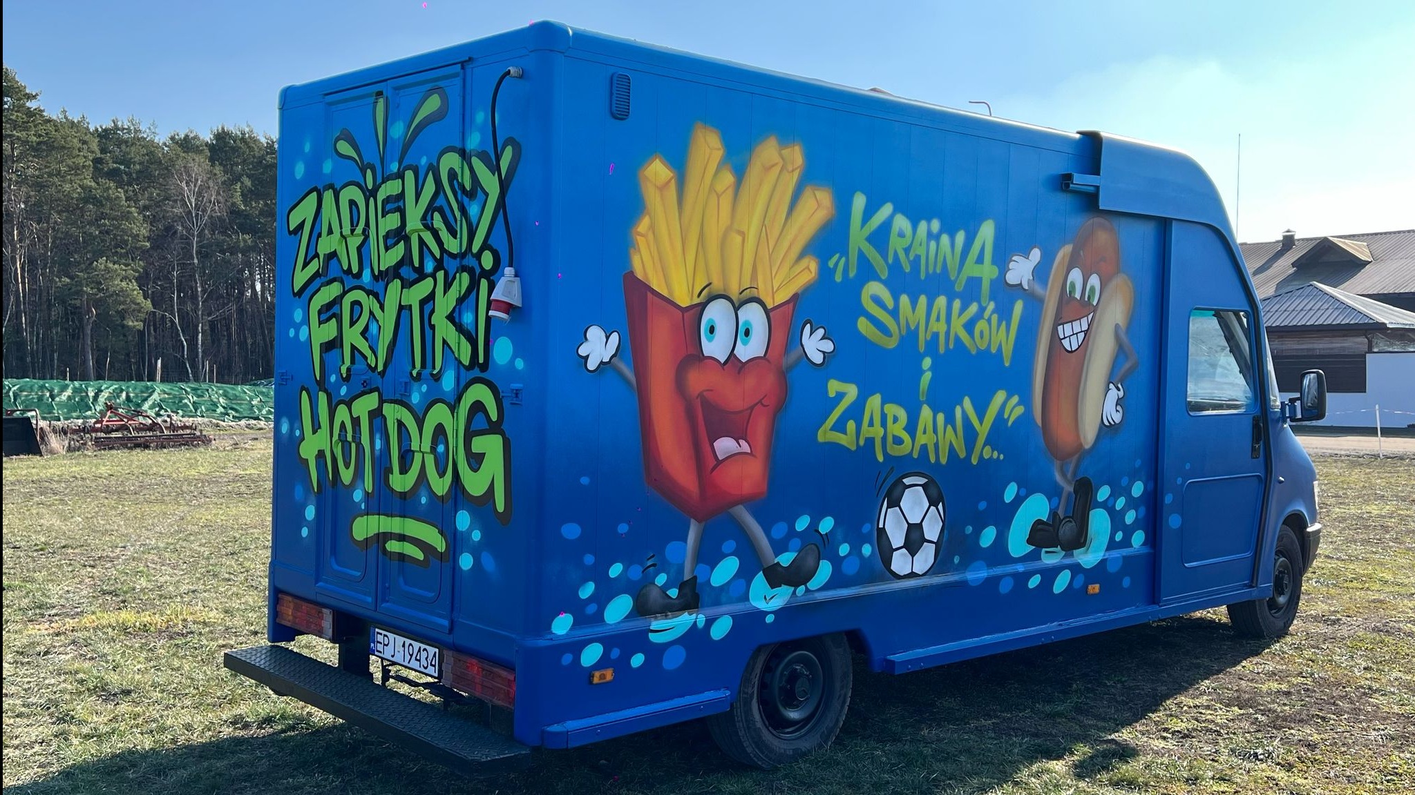 Niebieski food truck z kolorowymi malunkami frytek, hot dogów i napisem 'Kraina Smaków i Zabawy' na tle łąki i lasu.