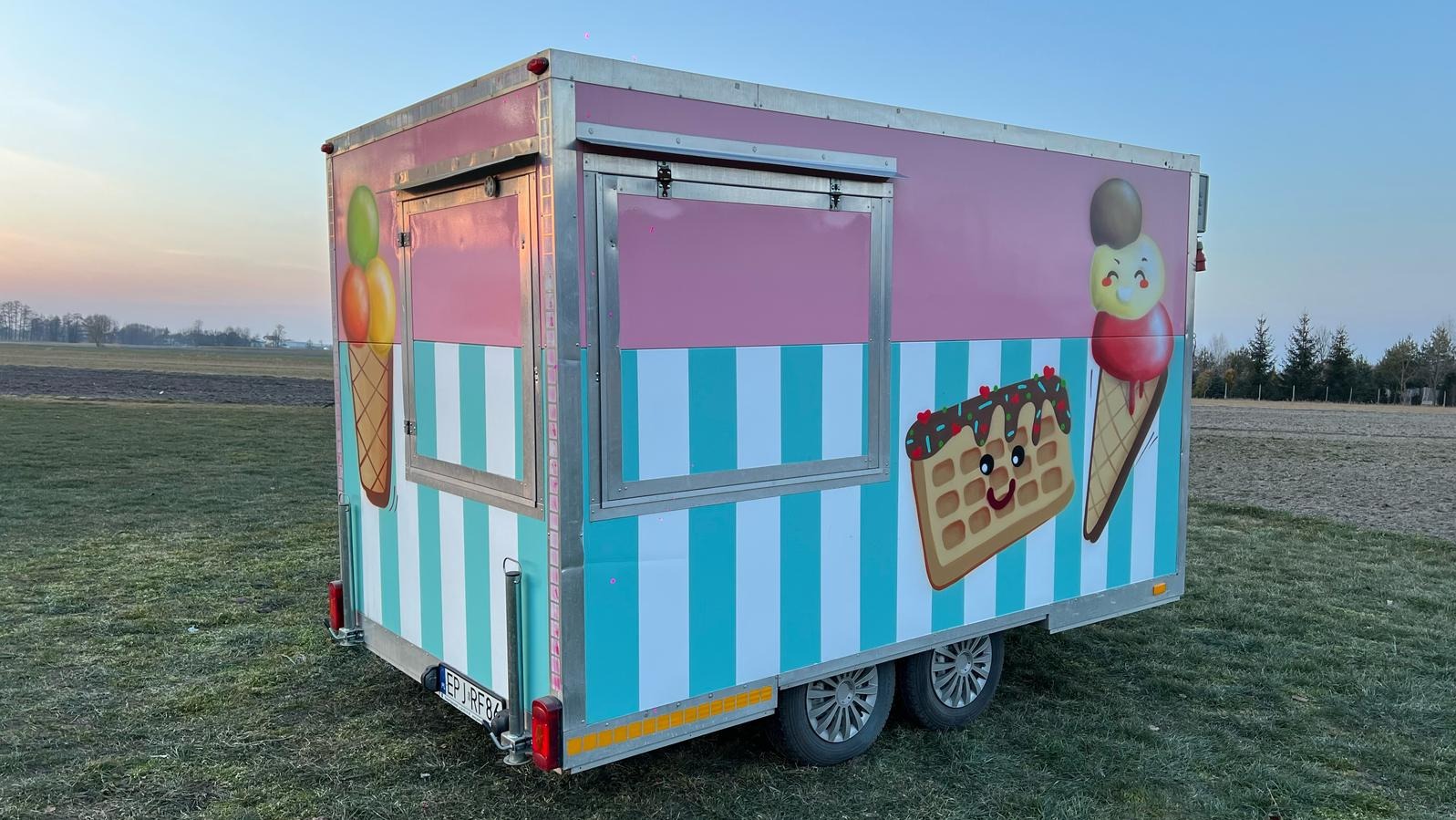 Przyczepa gastronomiczna z grafiką lodów i gofrów na tle łąki, z otwartym okienkiem sprzedażowym.