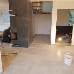 Usługi remontowo budowlane - Montaż obudowy kominka z szarych płyt imitujących marmur, widoczny pracownik podczas instalacji drzwiczek, surowy stan wykończenia wnętrza z widocznymi płytkami podłogowymi i przygotowanym otworem...