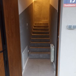Usługi remontowo budowlane - Wnętrze klatki schodowej z widocznymi schodami, grzejnikiem i oświetleniem, widziane zza otwartych drzwi wejściowych z numerem ulicy na ścianie.