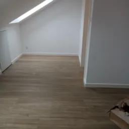 Poddasze po remoncie z oknem dachowym, świeżo ułożone panele podłogowe imitujące drewno, białe ściany i listwy przypodłogowe, karton po prawej stronie.