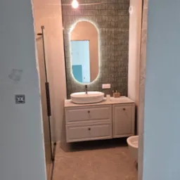 Nowoczesna łazienka z owalnym lustrem podświetlanym LED, białą umywalką nablatową i płytkami w odcieniach beżu i szarości, widziana z korytarza.