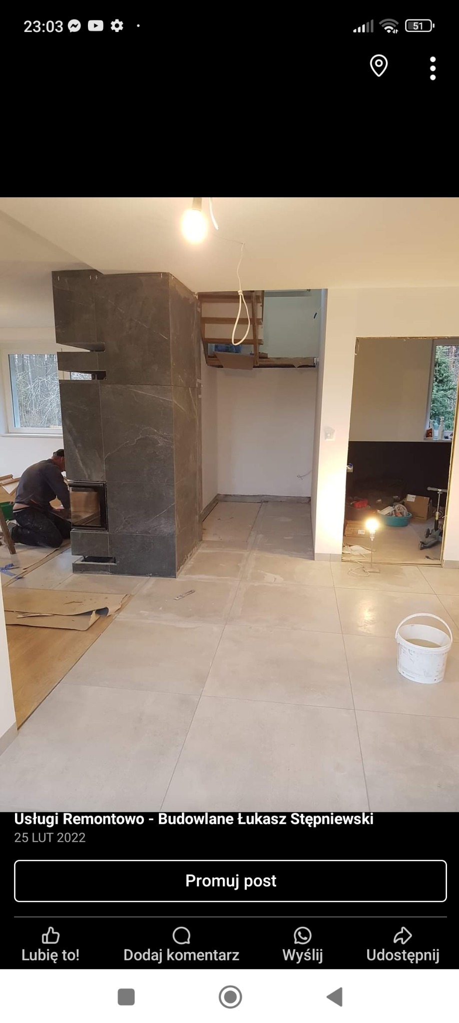 Montaż obudowy kominka z szarych płyt imitujących marmur, widoczny pracownik podczas instalacji drzwiczek, surowy stan wykończenia wnętrza z widocznymi płytkami podłogowymi i przygotowanym otworem...