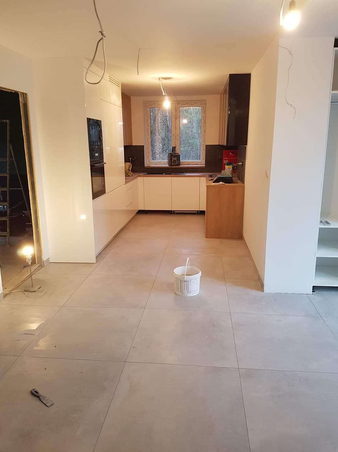 Wnętrze kuchni w trakcie prac wykończeniowych: białe, lakierowane szafki, szare płytki podłogowe, widoczne niedokończone ściany i sufit, wiadro po farbie na podłodze, drabina w tle.