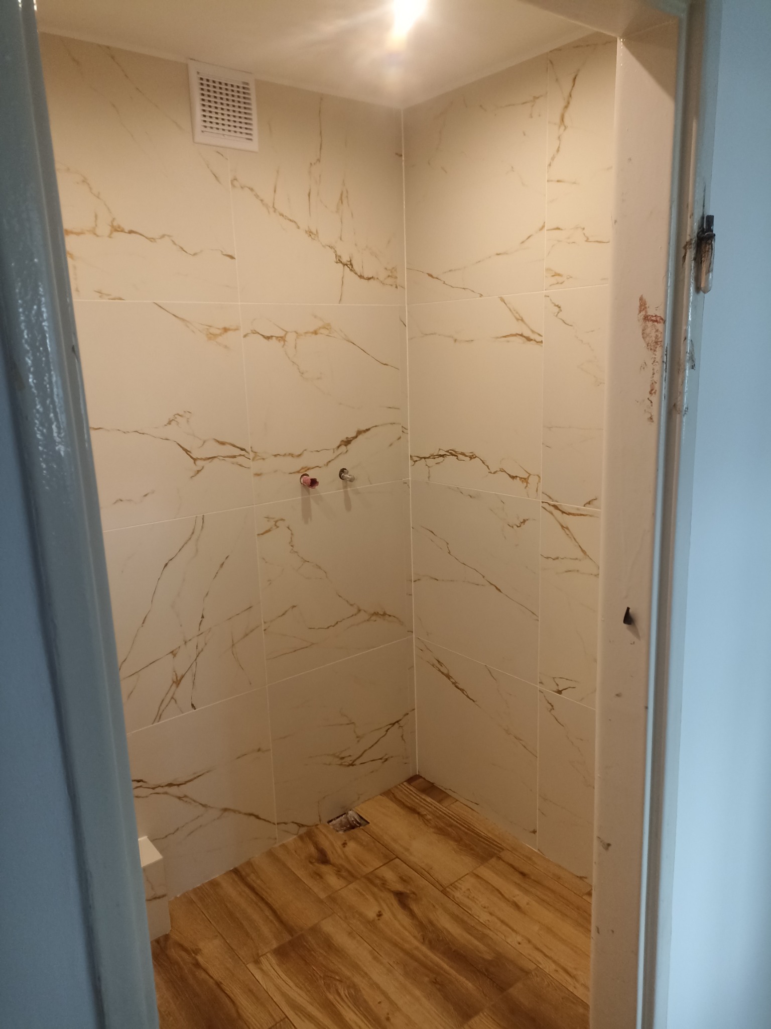 Wykończona łazienka z beżowymi płytkami imitującymi marmur na ścianach i drewnopodobną podłogą, widok z zewnątrz przez drzwi.