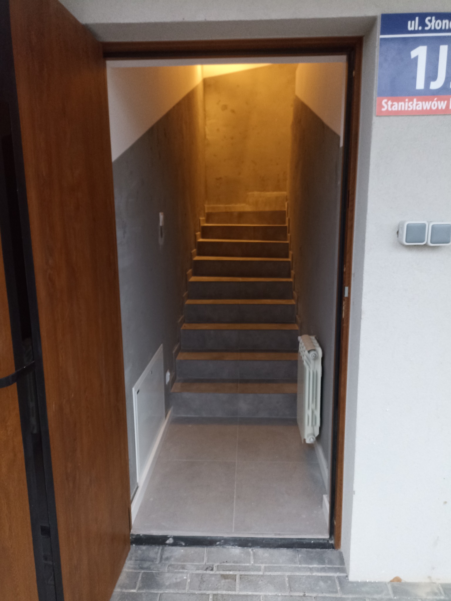 Wnętrze klatki schodowej z widocznymi schodami, grzejnikiem i oświetleniem, widziane zza otwartych drzwi wejściowych z numerem ulicy na ścianie.