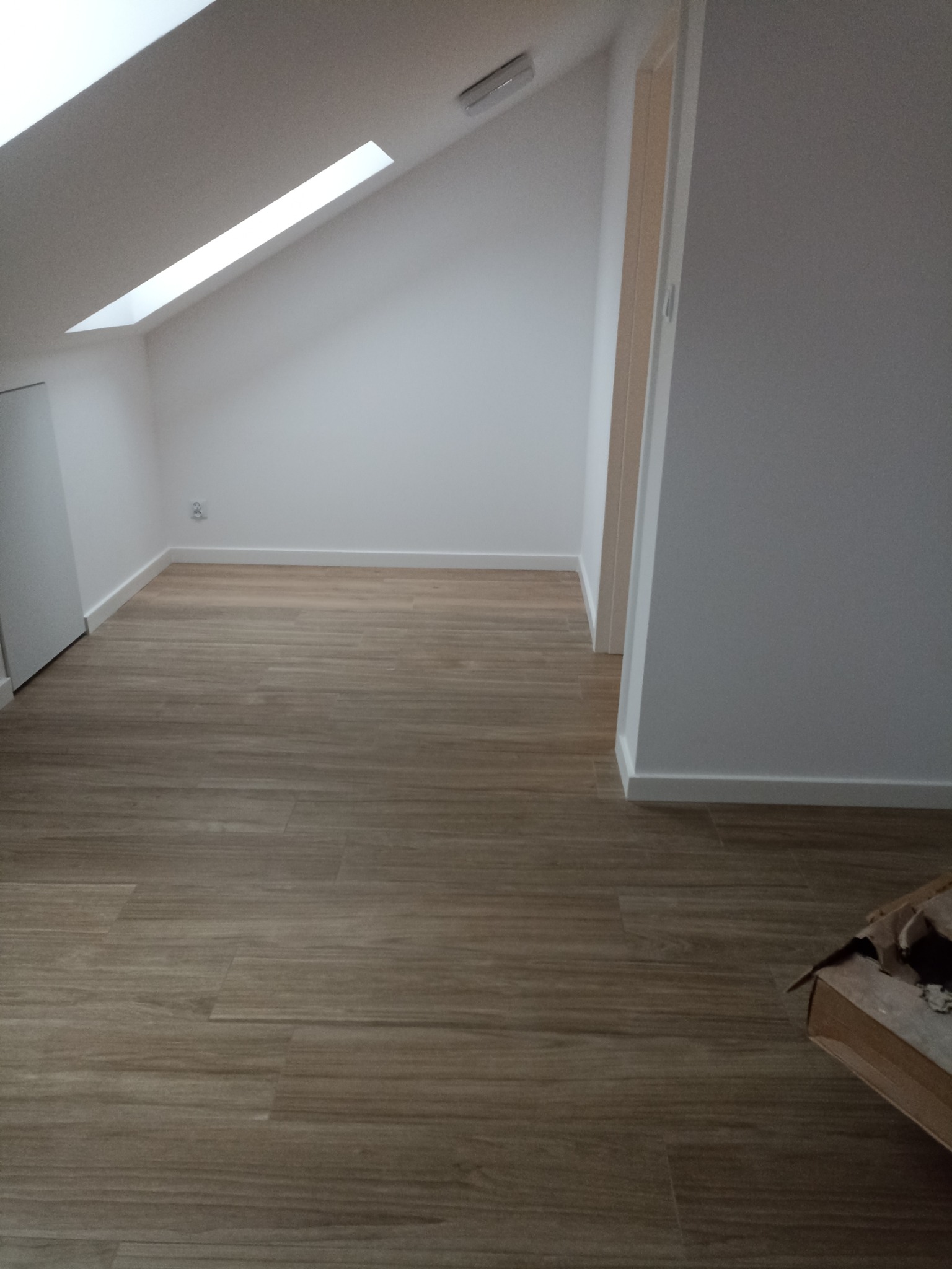 Poddasze po remoncie z oknem dachowym, świeżo ułożone panele podłogowe imitujące drewno, białe ściany i listwy przypodłogowe, karton po prawej stronie.