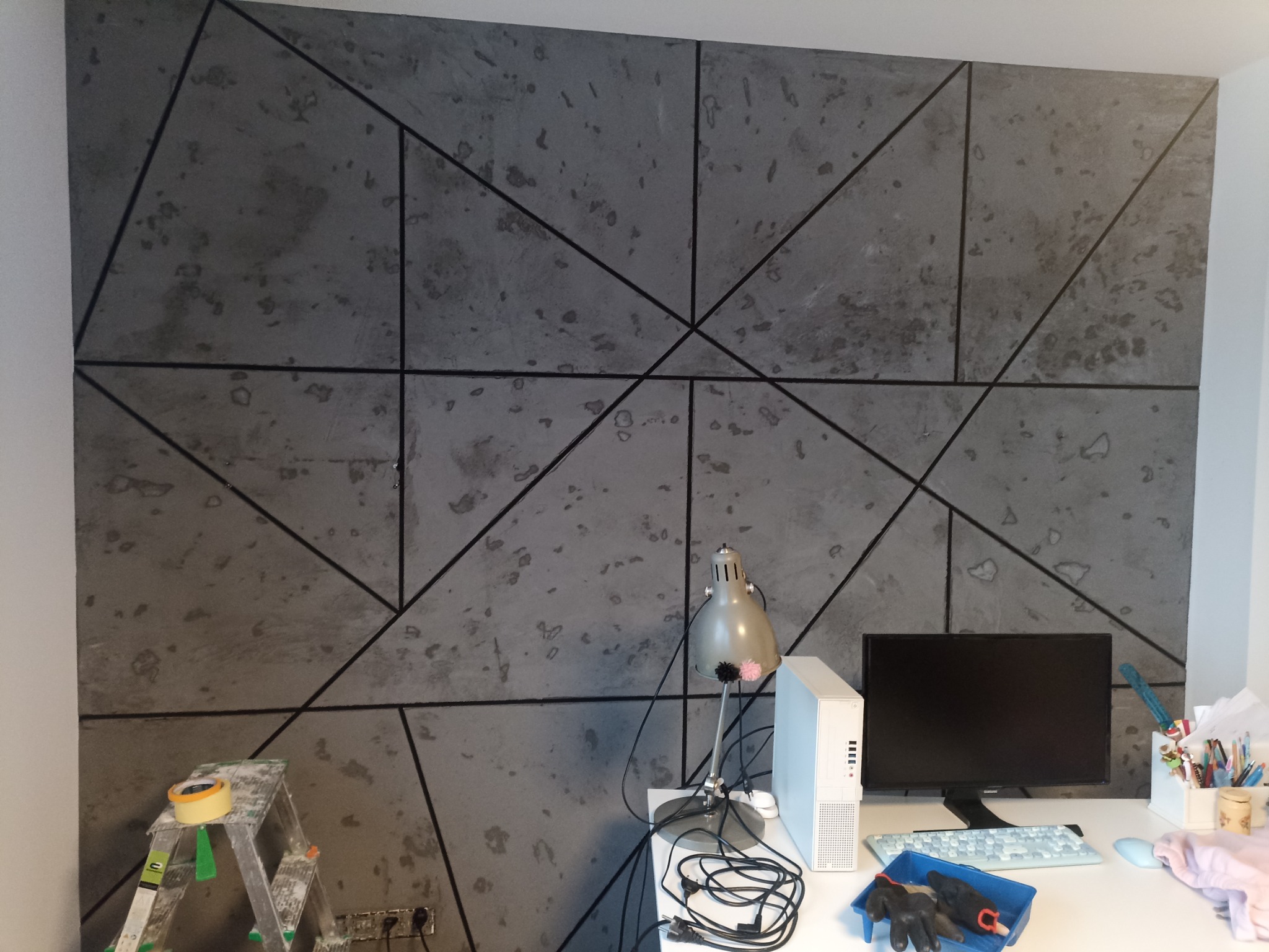 Dekoracyjna ściana w pokoju z szarego betonu architektonicznego z geometrycznym podziałem czarnymi liniami, widoczny fragment biurka z komputerem i drabina malarska z taśmą maskującą.