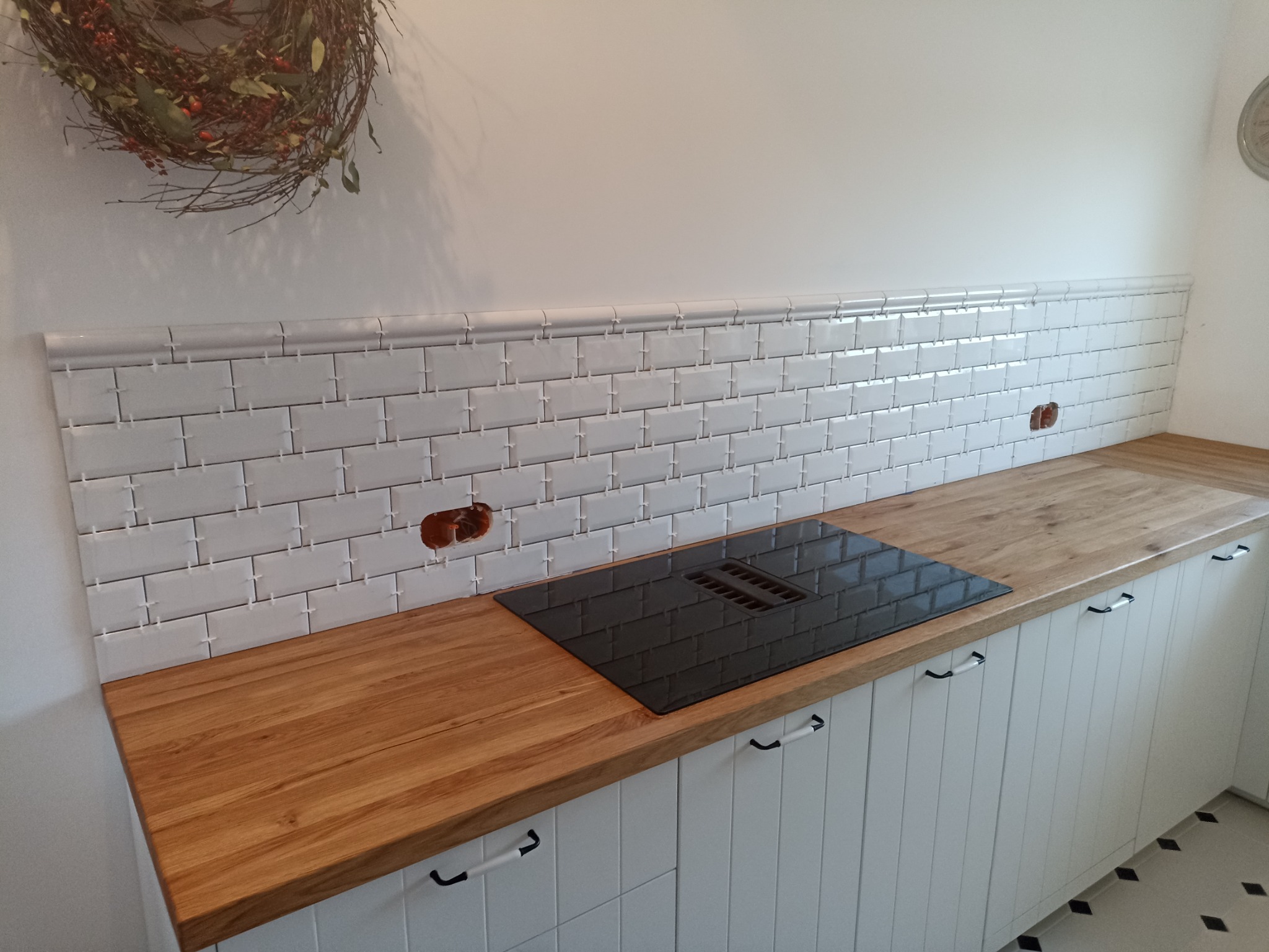 Kuchnia w stylu retro z drewnianym blatem, białymi szafkami z czarnymi uchwytami i białymi płytkami ułożonymi na ścianie, z widocznymi gniazdkami elektrycznymi i czarną płytą grzewczą...