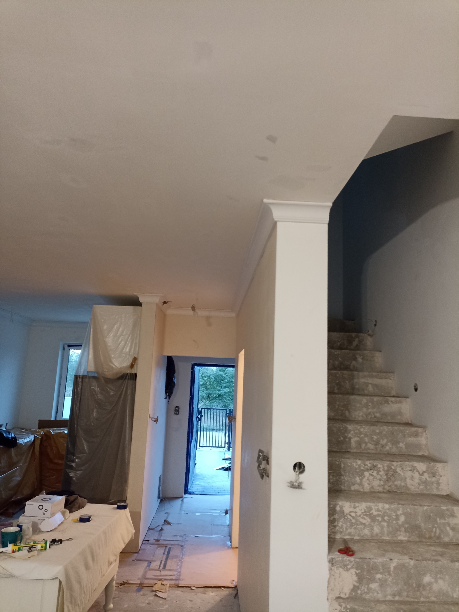 Wnętrze w trakcie prac: surowe schody obok kolumny z listwą dekoracyjną, zabezpieczone meble folią, widoczne ślady po szpachlowaniu na suficie, przygotowane narzędzia na stole.
