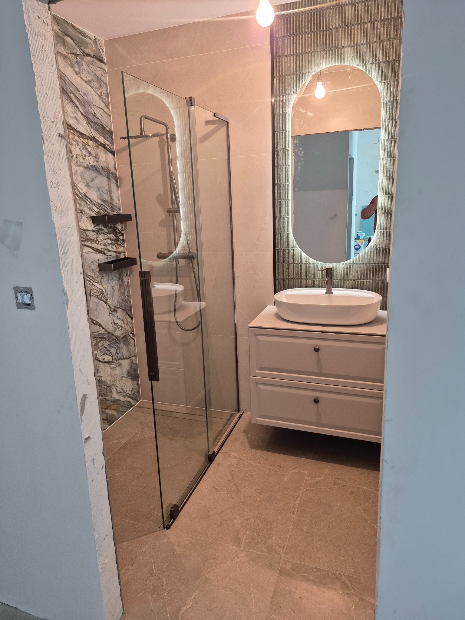 Nowoczesna łazienka w trakcie wykończenia: kabina prysznicowa ze szklanymi drzwiami, umywalka na szafce z lustrem z podświetleniem LED. Widoczne niedokończone ściany.